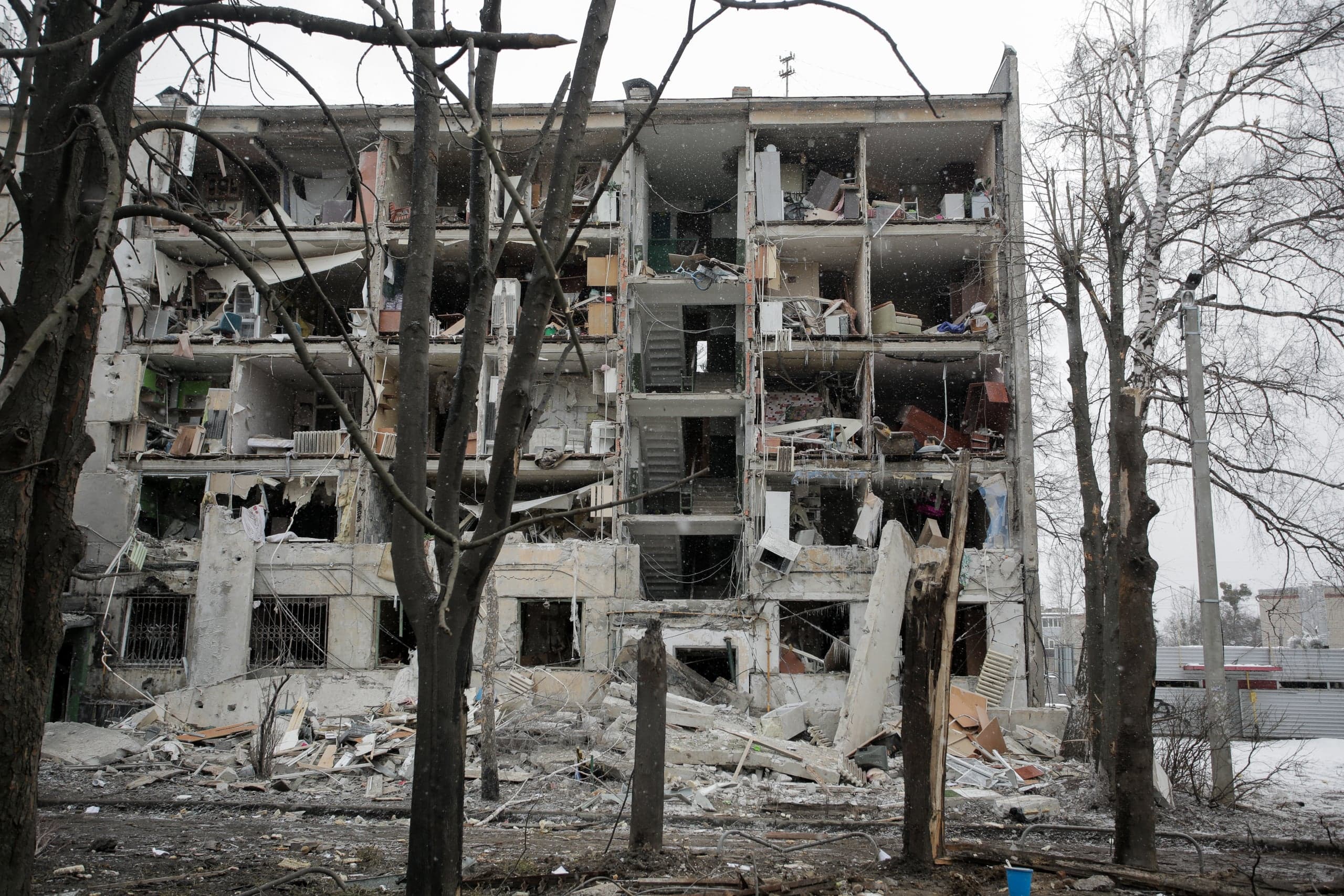 Сильно разрушенный обстрелом многоэтажный жилой дом в снежный день. Центральная часть здания уничтожена, видны внутренние помещения с обломками. На переднем плане — голые деревья и строительный мусор.