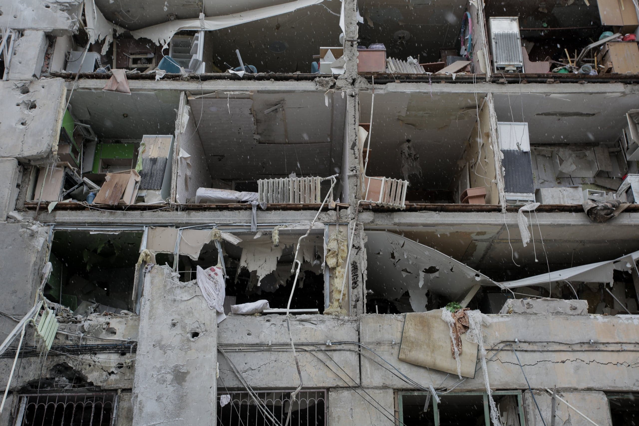 Крупный план фасада разрушенного обстрелом жилого дома, где видны открытые секции трех этажей с остатками мебели и падающим снегом.