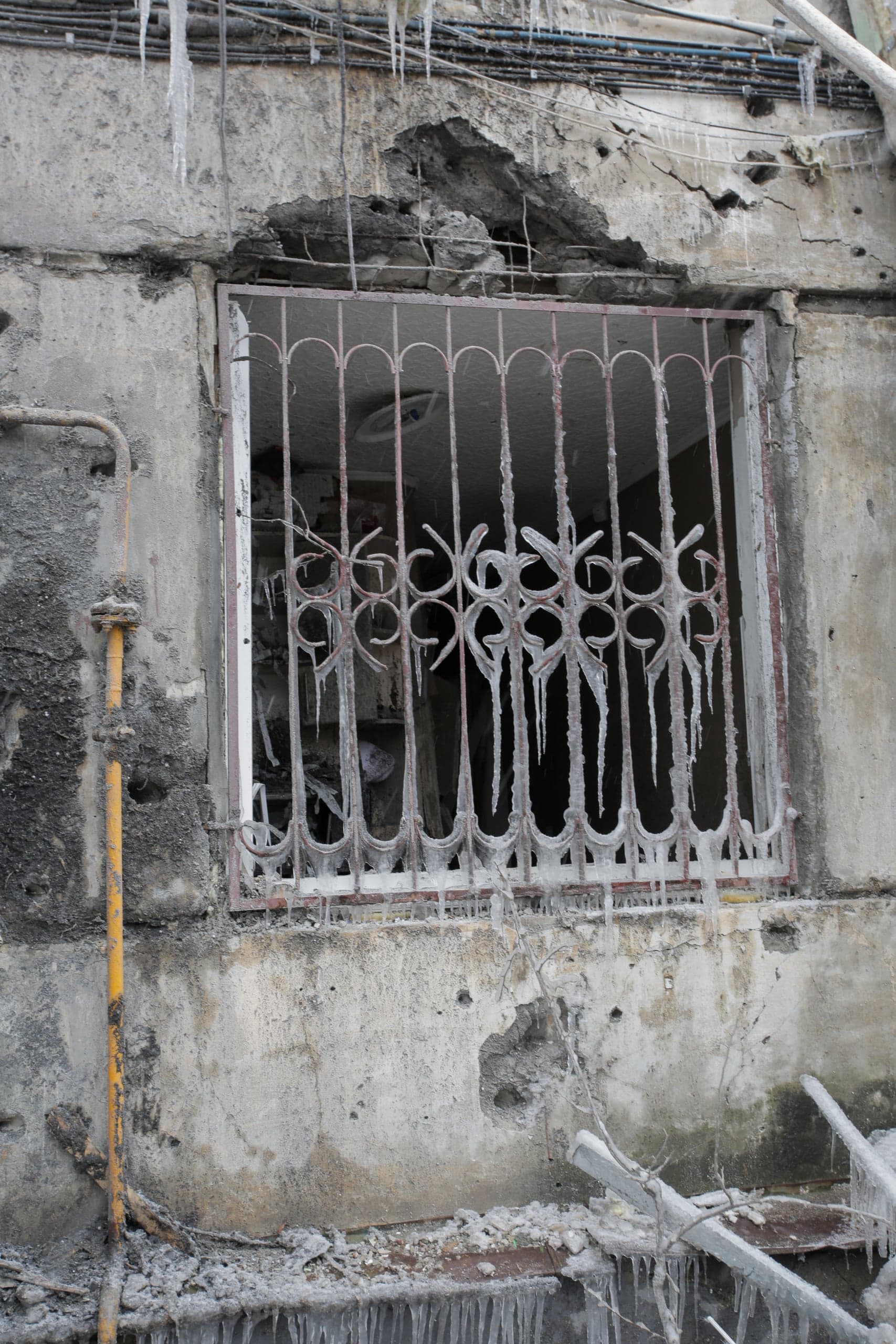 Поврежденная бетонная стена с разрушенным оконным проемом. Металлическая решетка окна и стены густо покрыты сосульками и льдом.