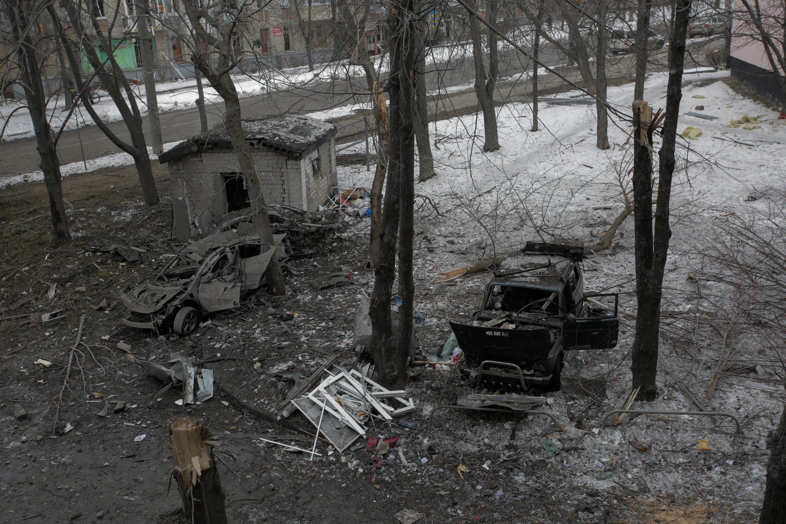 Разрушенный городской пейзаж зимой или ранней весной. На грязном, частично заснеженном дворе лежат обломки двух уничтоженных автомобилей, руины небольшого кирпичного строения, груды мусора и сломанные стволы деревьев.
