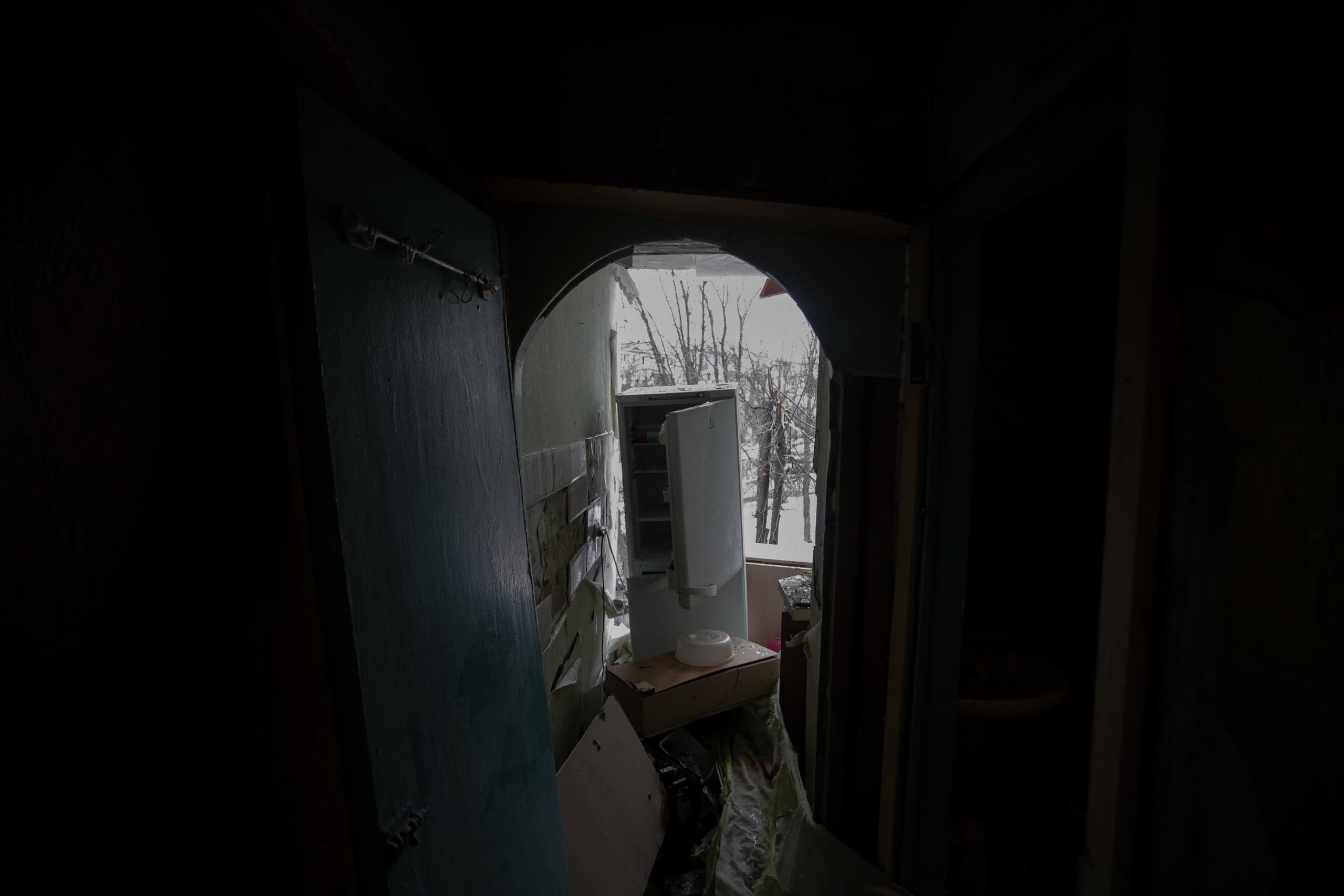 Вид из темного проема в разрушенную комнату. В центре стоит белый холодильник; за ним через большой пролом виден заснеженный зимний пейзаж с голыми деревьями.