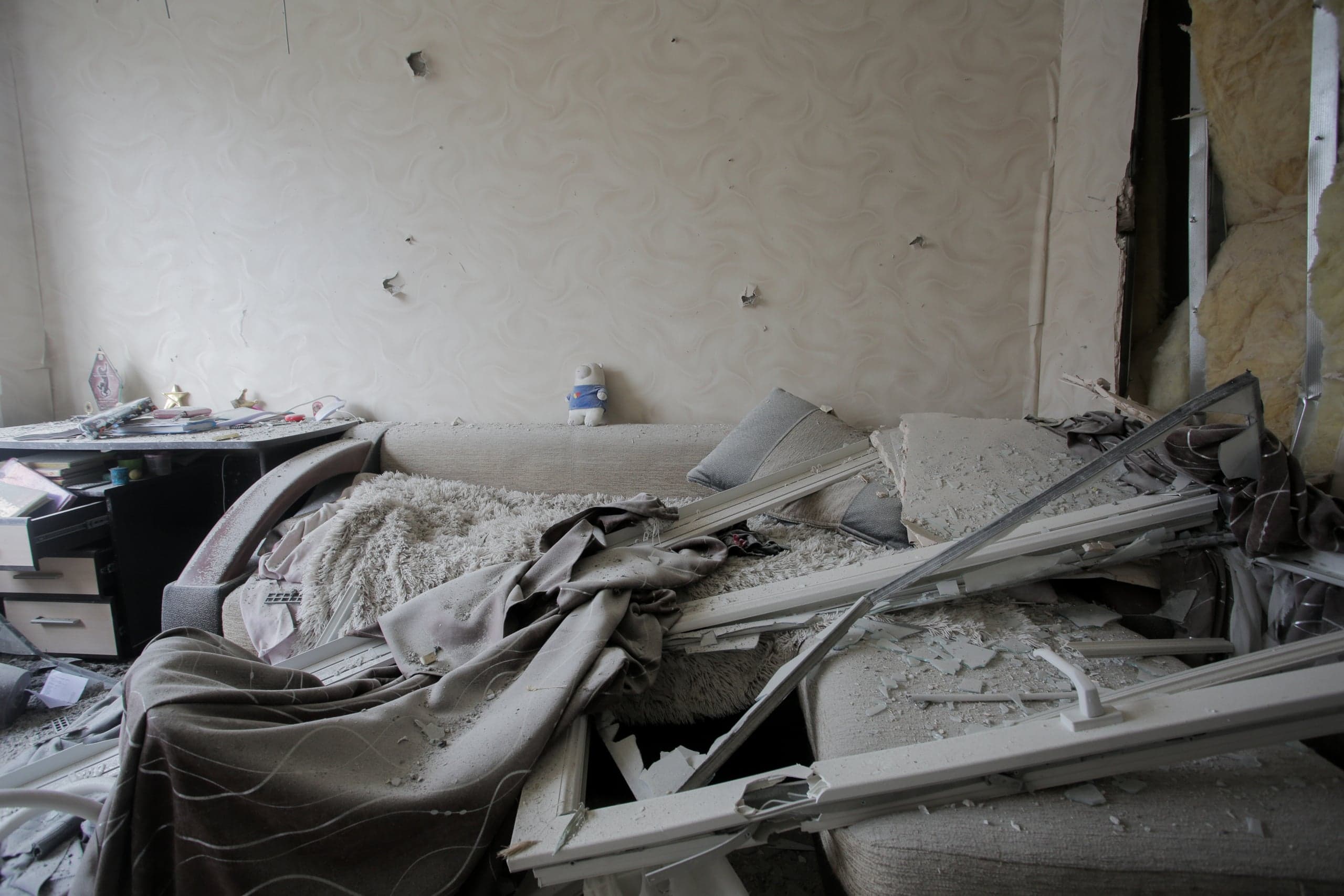 Интерьер жилой комнаты, разрушенной в результате обстрела: обломки белых оконных рам, осколки стекла и пыль покрывают диван. На стене видны многочисленные отверстия от осколков; на спинке дивана стоит мягкая игрушка в синей футболке.