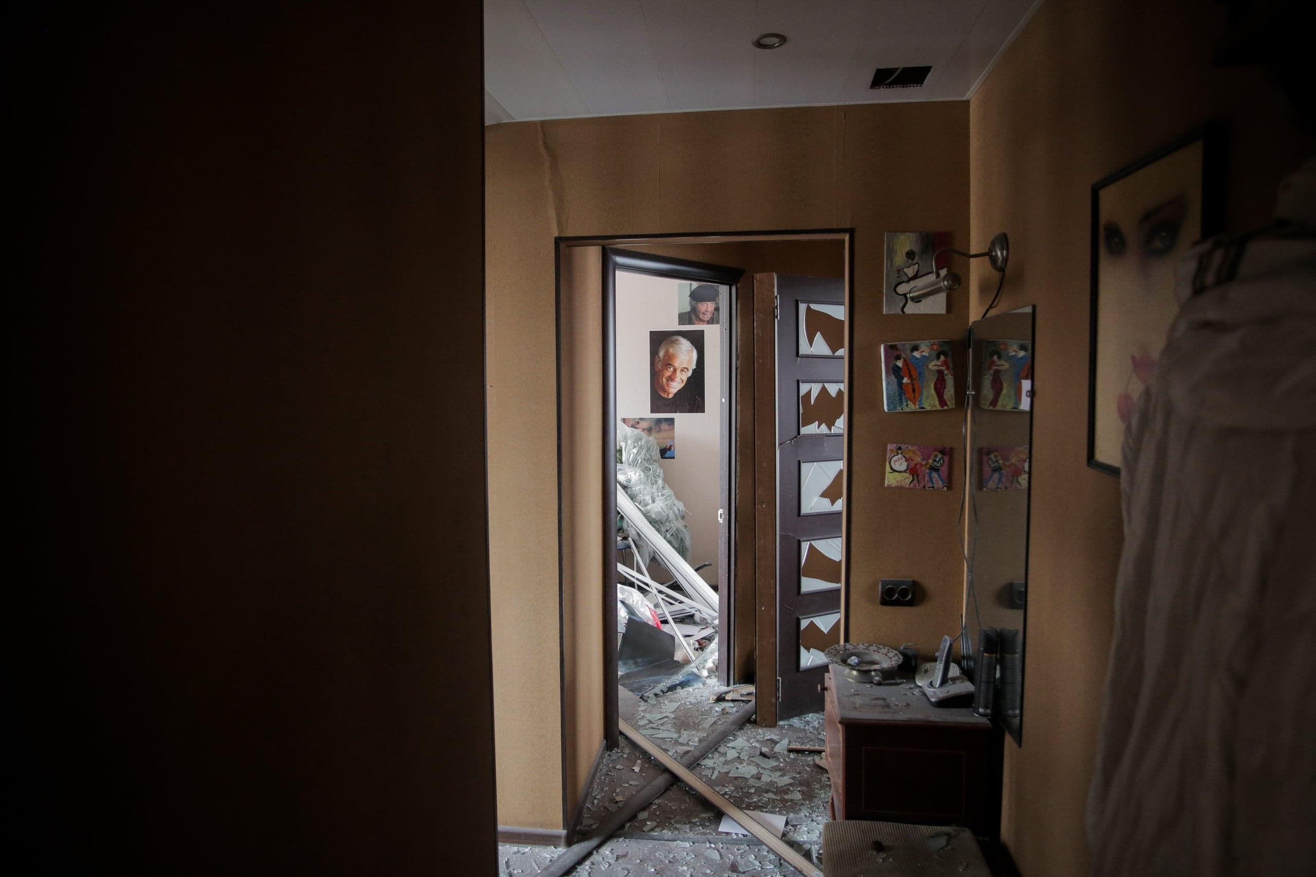 Коридор поврежденной квартиры, пол которого усеян осколками стекла и обломками. Открытый дверной проем ведет в сильно разрушенную комнату; на стенах коридора висят фотографии и картины.
