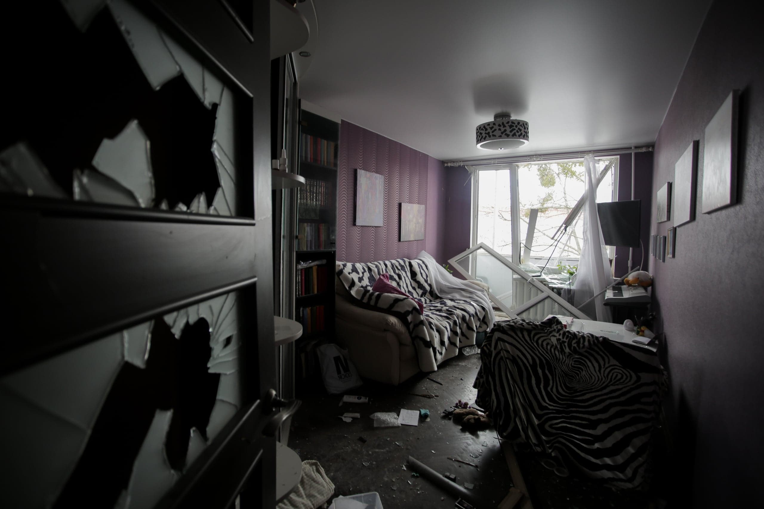 Поврежденная жилая комната, залитая светом из полностью разбитого окна, рама которого лежит на полу. На переднем плане — крупным планом разбитое стекло в дверном проеме. Пол завален осколками и мусором.