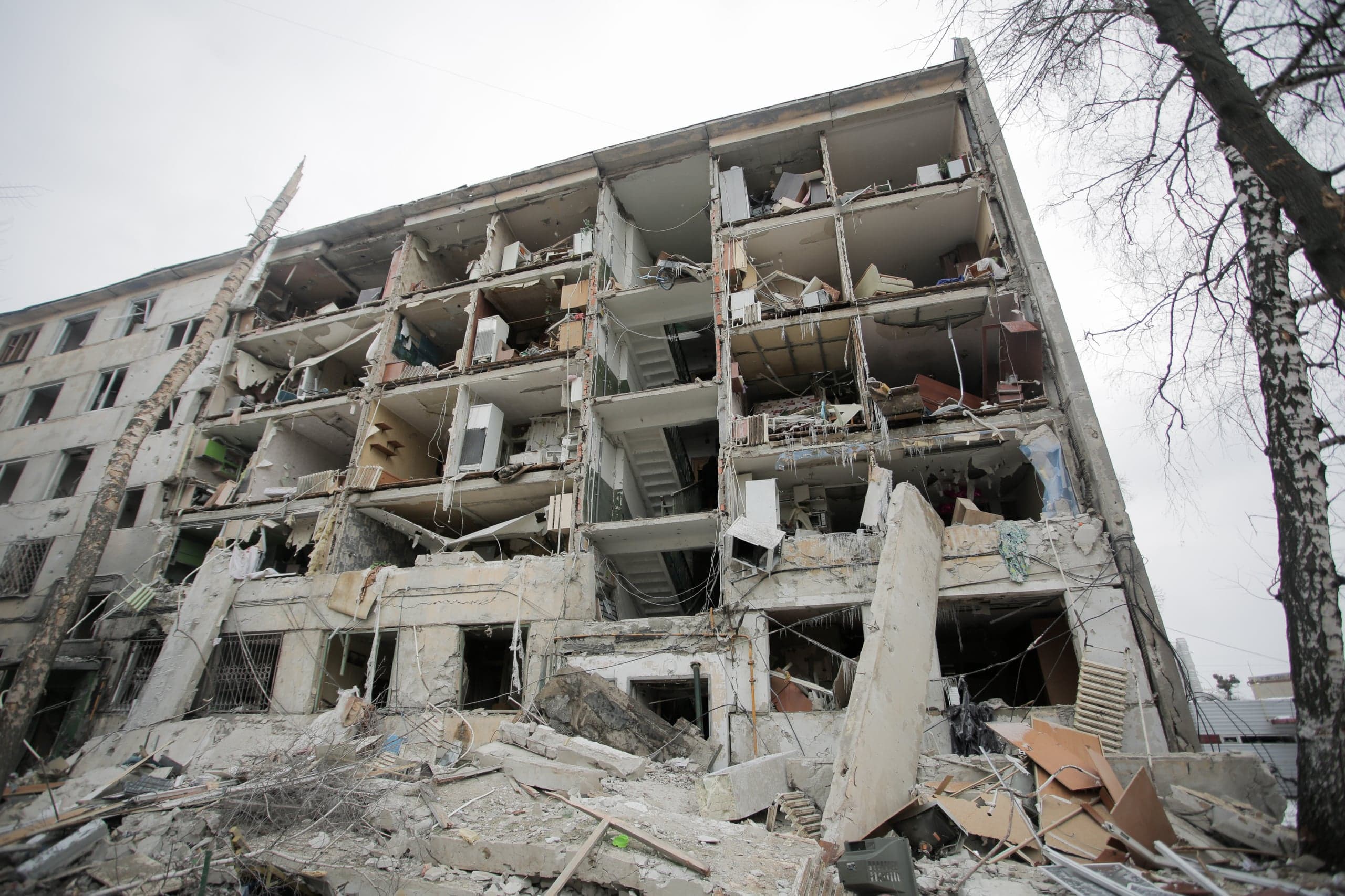 Сильно разрушенный взрывом многоквартирный дом. Обнажены этажи, лестничные пролеты и остатки мебели, а перед зданием лежит большая груда бетонных обломков. Справа видны деревья.