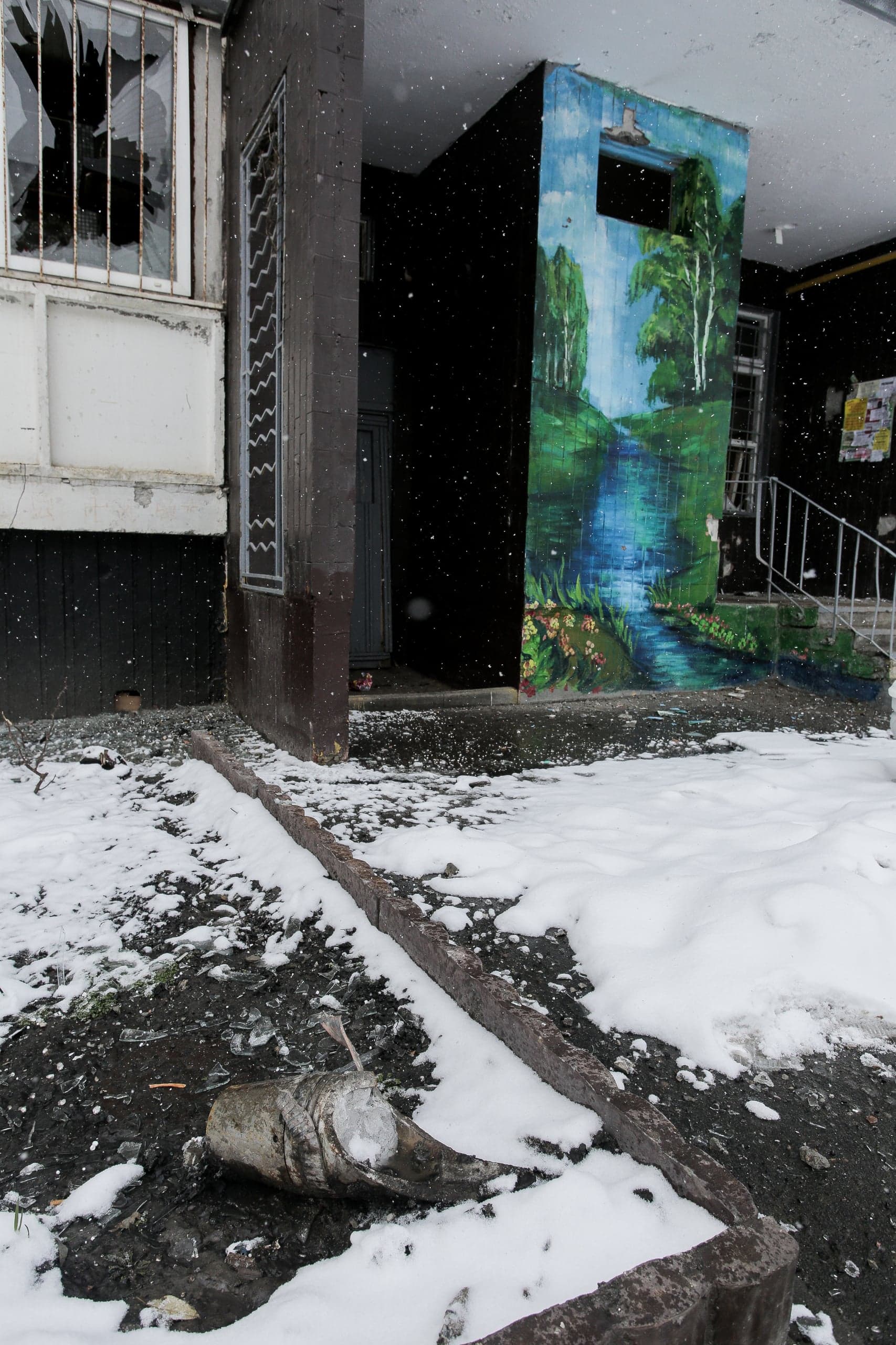 Вход в поврежденное здание со сломанным окном и фреской. На заснеженной земле лежит крупный фрагмент боеприпаса.