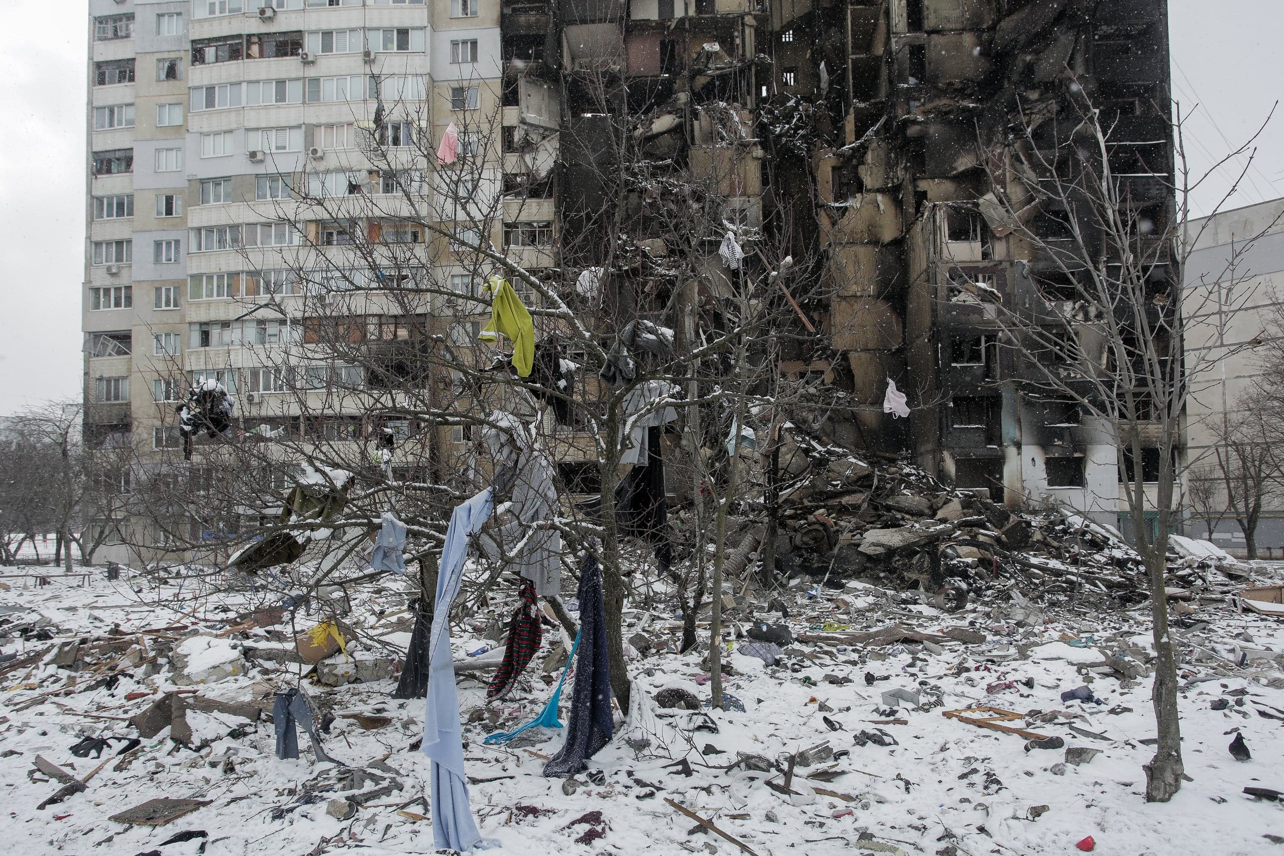 Разрушенный и обгоревший многоэтажный дом зимой; на заснеженных деревьях и земле разбросана одежда и обломки.