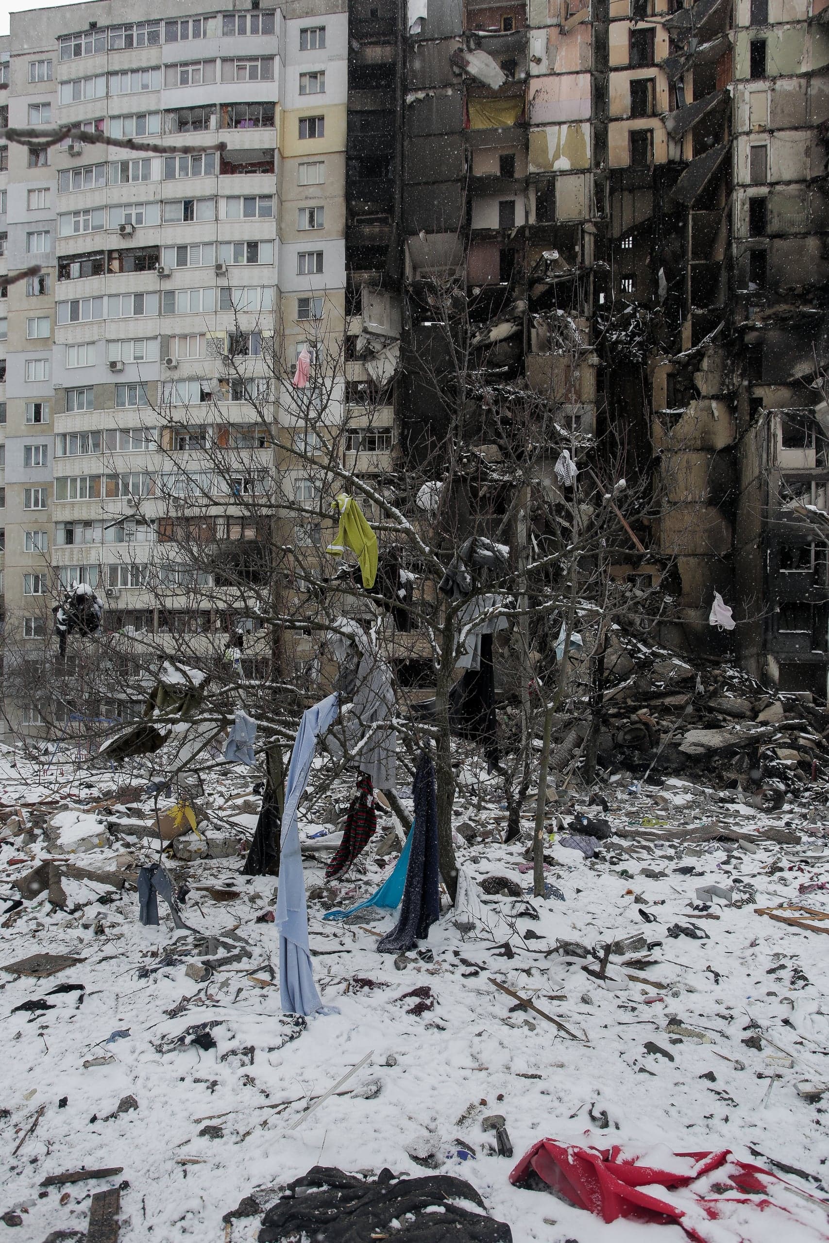 Сгоревший и частично разрушенный многоэтажный дом на фоне снега. Одежда висит на голых ветках деревьев среди обломков.