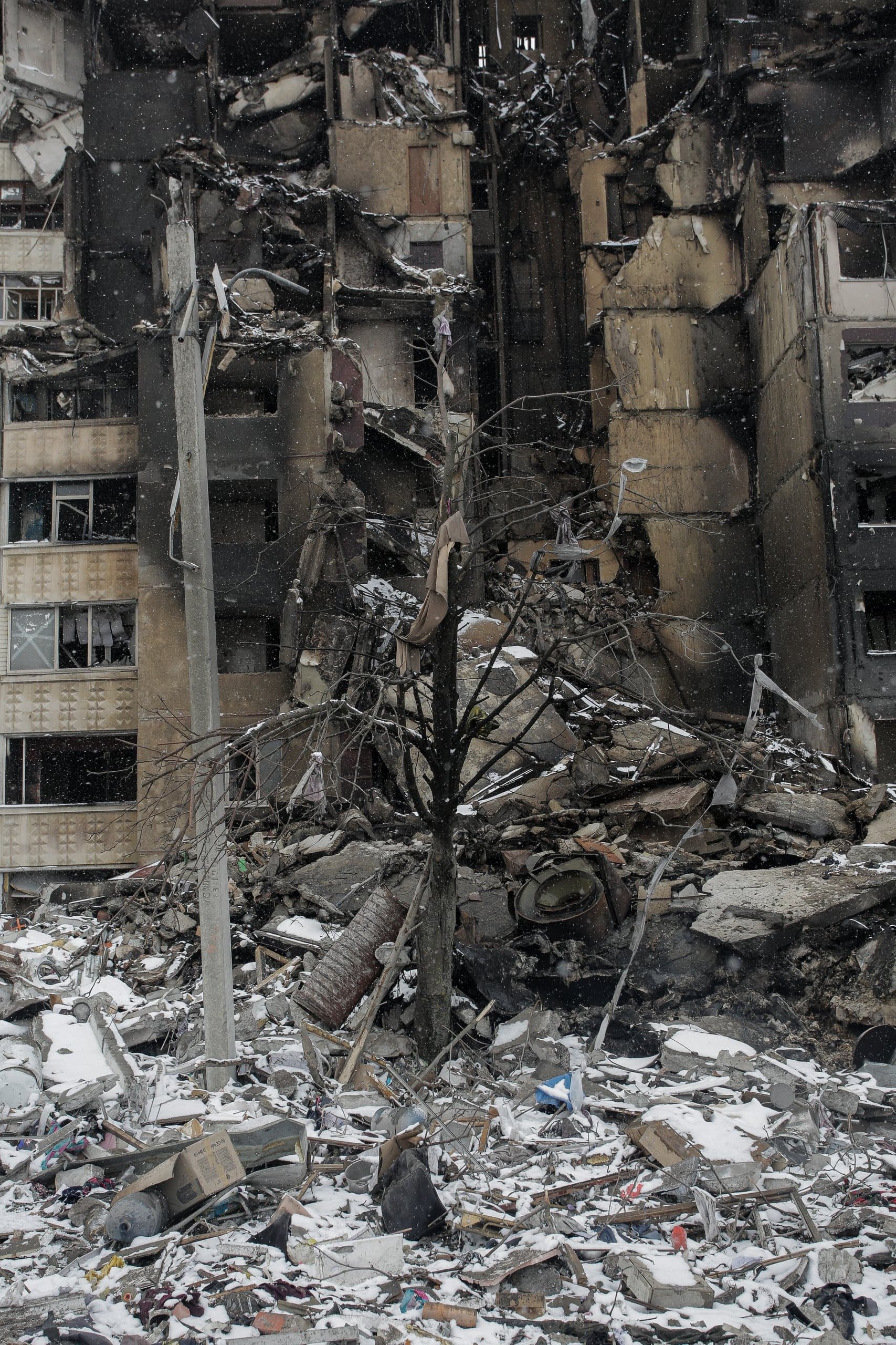 Разрушенное и обгоревшее многоэтажное здание, обломки и завалы на переднем плане под снегом.