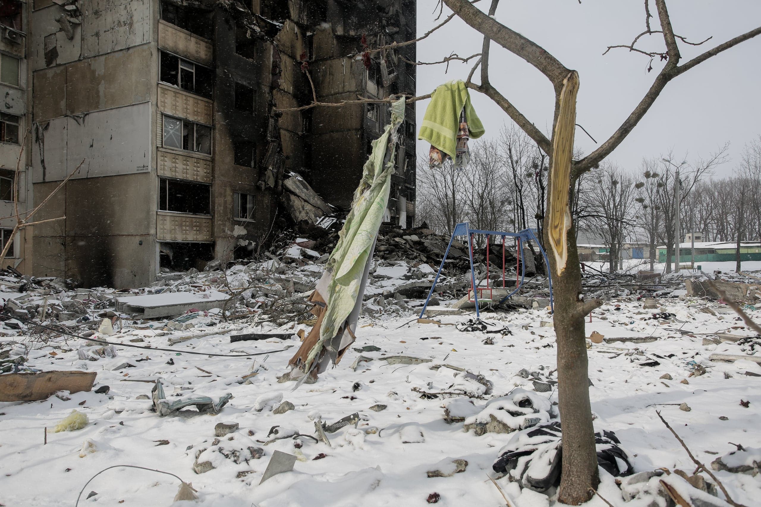 Обгоревший, частично разрушенный многоэтажный дом, заснеженный двор, усеянный обломками. На ветке сломанного дерева висит зеленое полотенце.