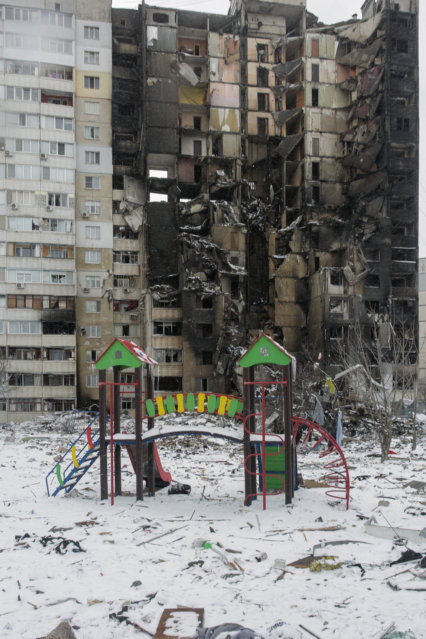 Обгоревший, частично разрушенный многоэтажный жилой дом. На переднем плане — заснеженная детская площадка, заваленная обломками.
