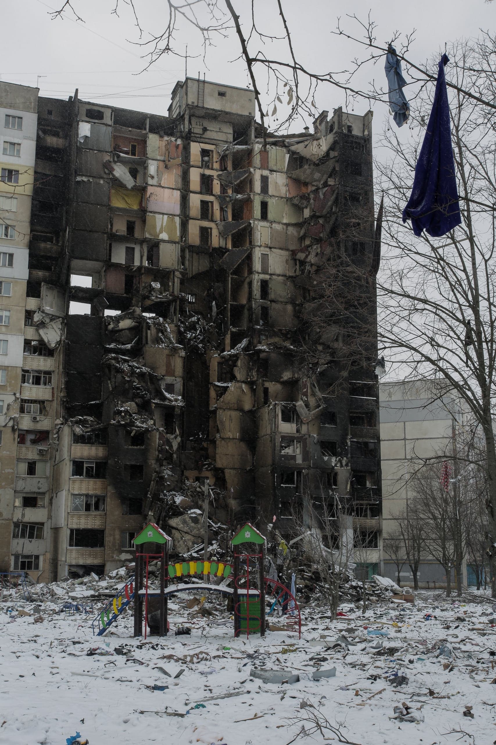 Разрушенный, обгоревший многоэтажный дом на фоне заснеженной, усыпанной обломками детской площадки.