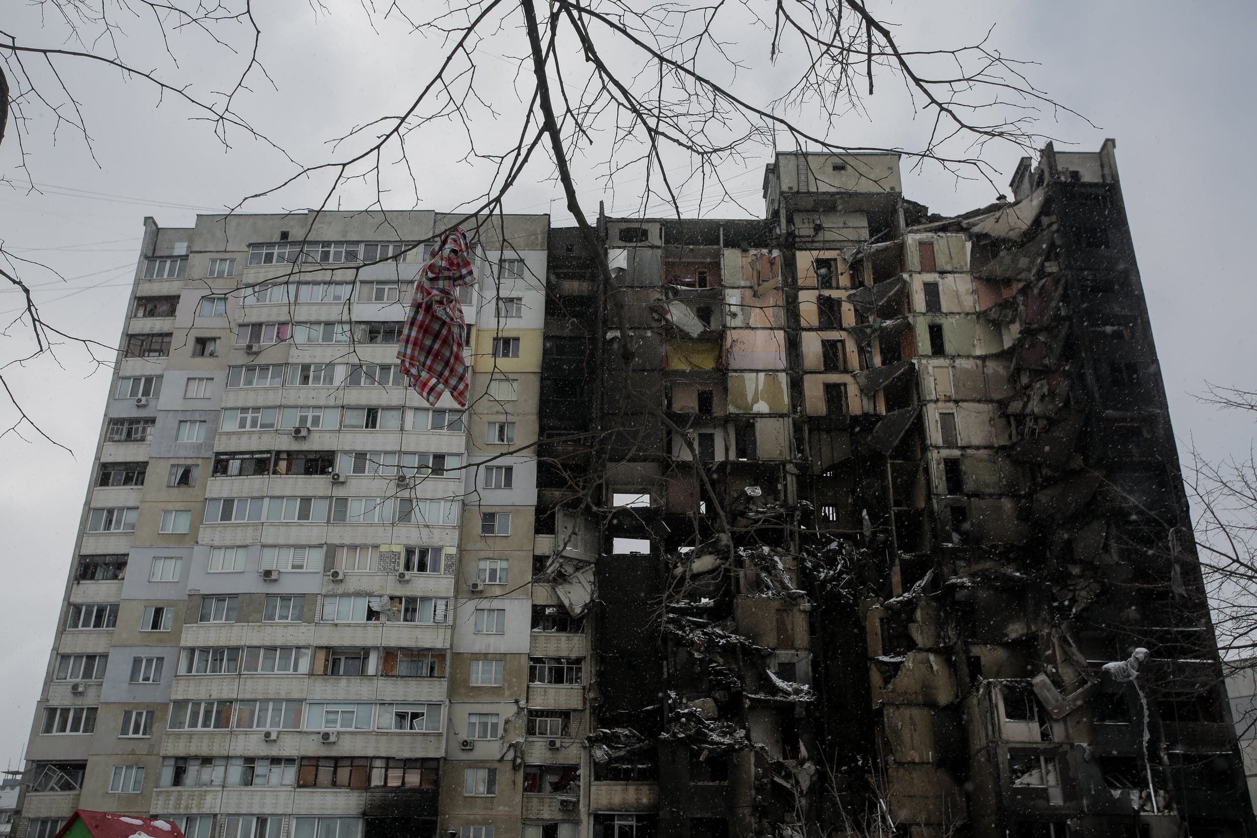 Поврежденный многоэтажный жилой дом: левая часть цела, правая обгоревшая и частично разрушенная. С ветки свисает красно-белая клетчатая ткань.