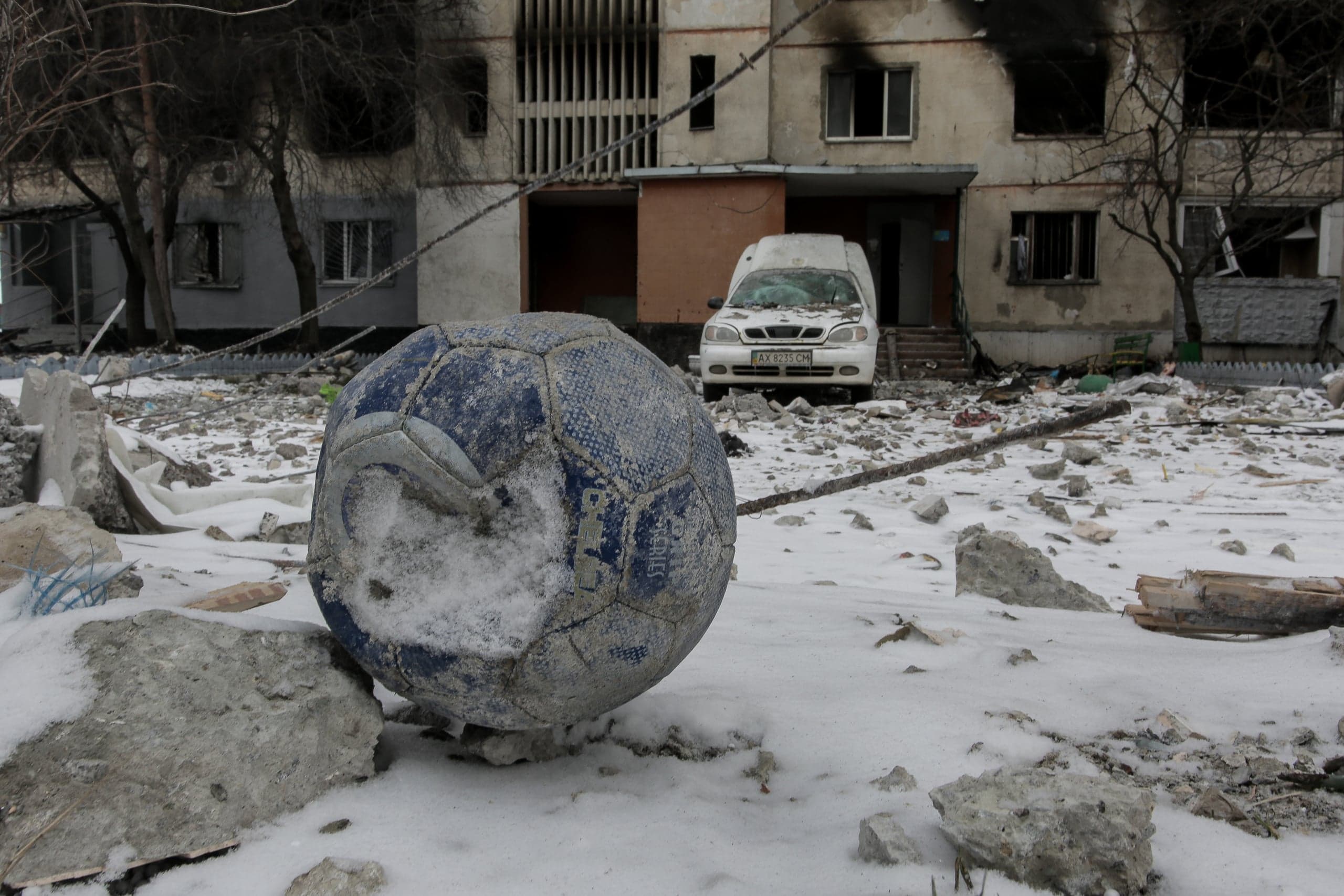 Сдутый футбольный мяч на снегу среди обломков; на фоне – разрушенный дом и поврежденный автомобиль с номером "AX 8215 CH".