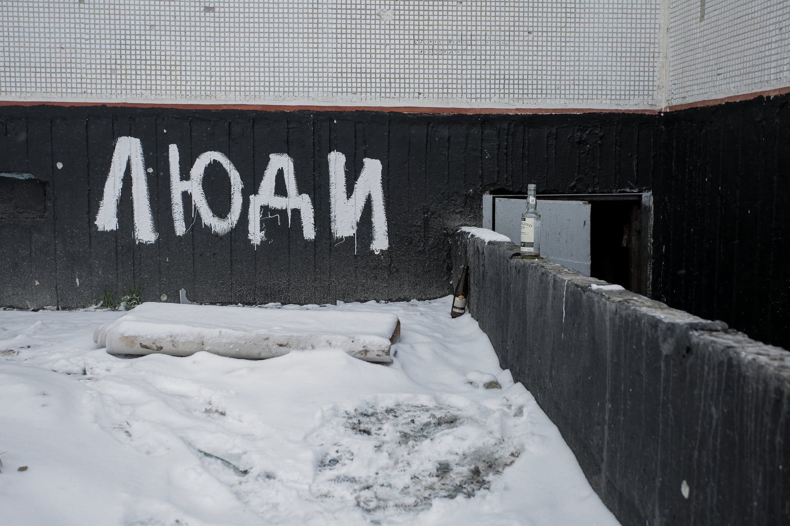 Снежный двор. На черной стене белое граффити "ЛЮДИ". На бетонном бордюре стоит пустая бутылка, на снегу лежит матрас.