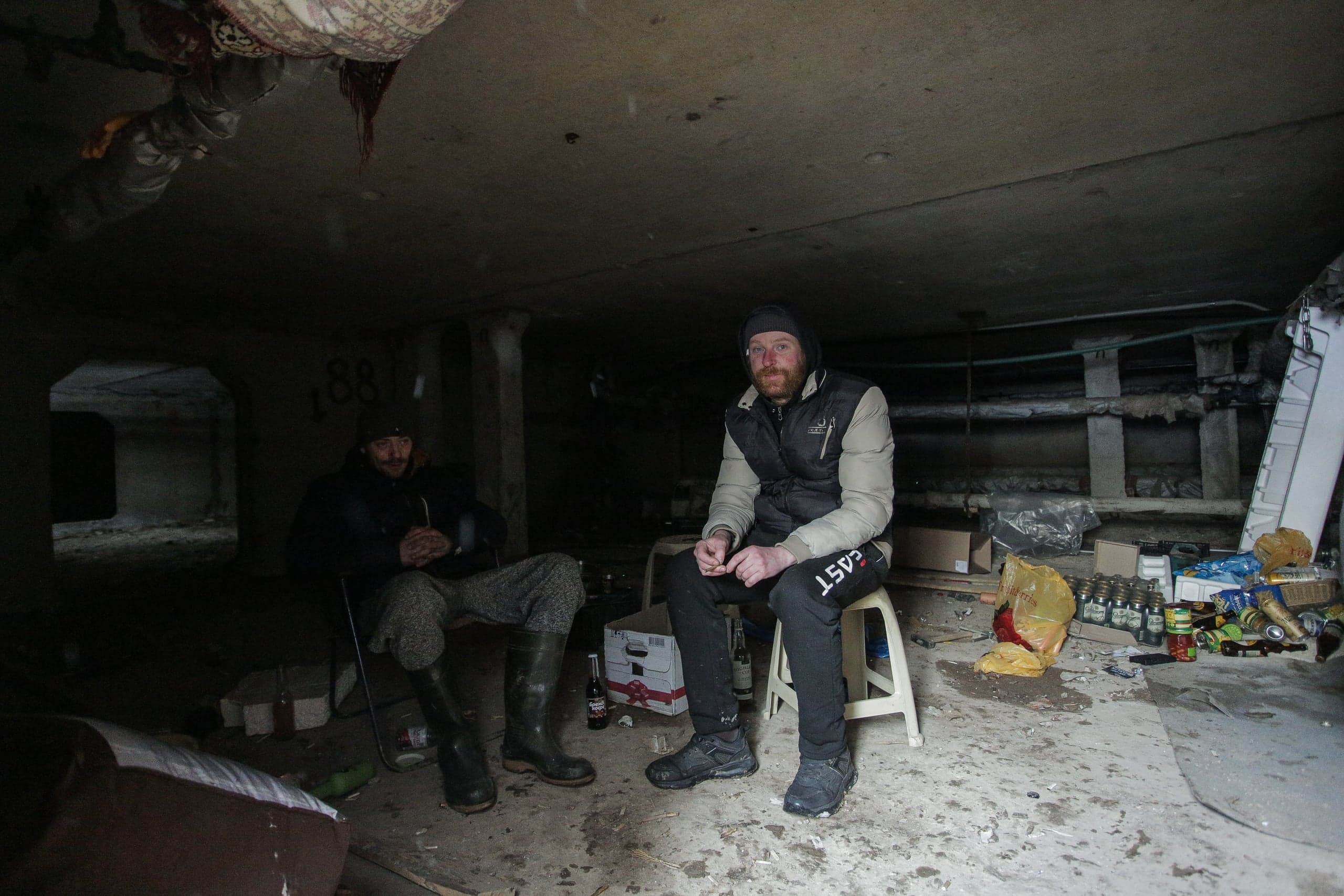 Двое мужчин сидят в тусклом, захламленном бетонном подземелье.
