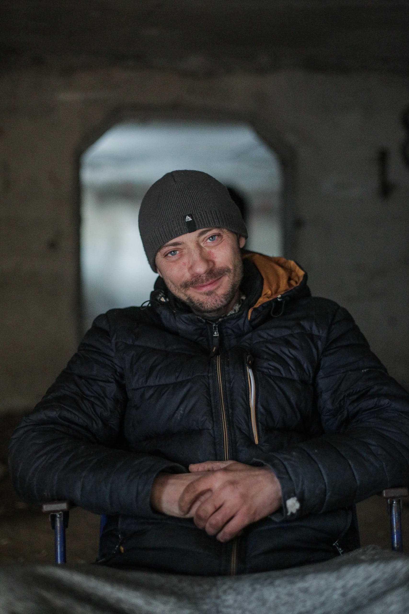 Мужчина в серой шапке и черной куртке сидит и улыбается в полутемном помещении.