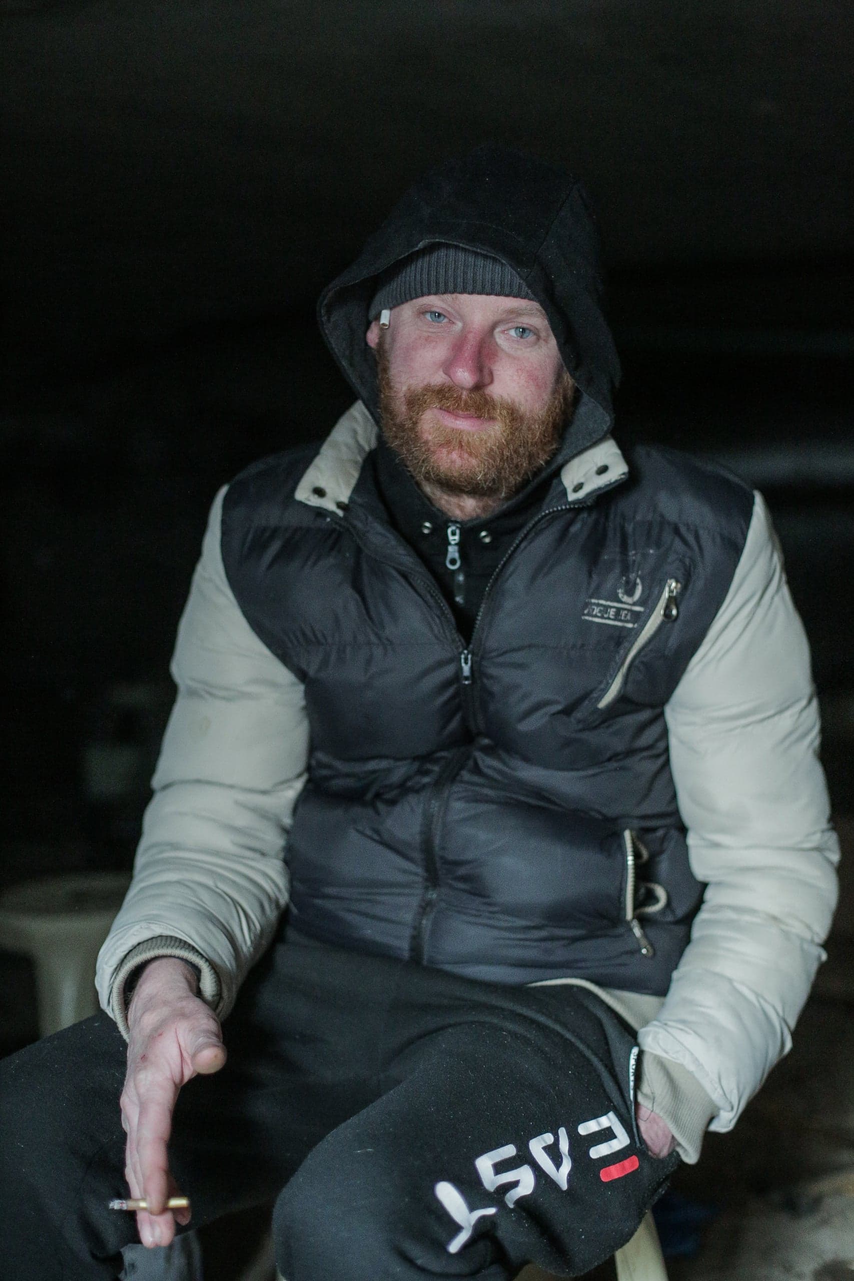 Рыжебородый мужчина в капюшоне, жилете и шапке сидит в темноте и держит сигарету. На спортивных штанах надпись "EAST".