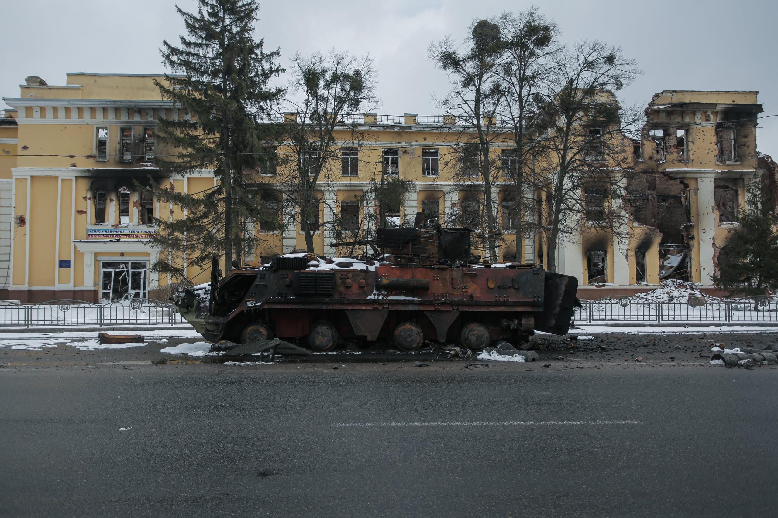 Обгоревший бронетранспортер на дороге перед сильно поврежденным, частично разрушенным желтым зданием. Зимний пейзаж.