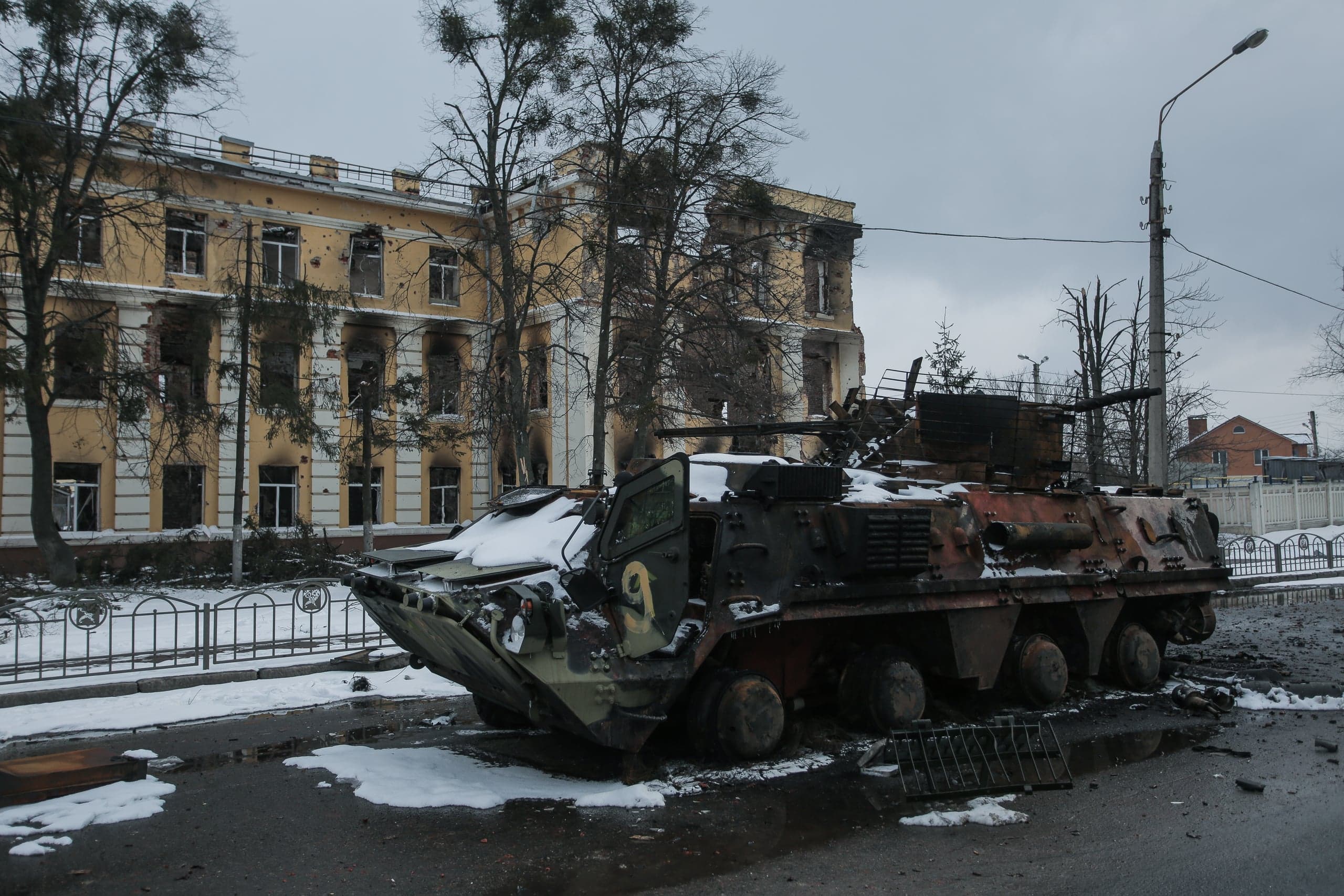 Обгоревший, подбитый бронетранспортер с цифрой "9" на заснеженной улице перед сильно разрушенным обстрелами желтым зданием.