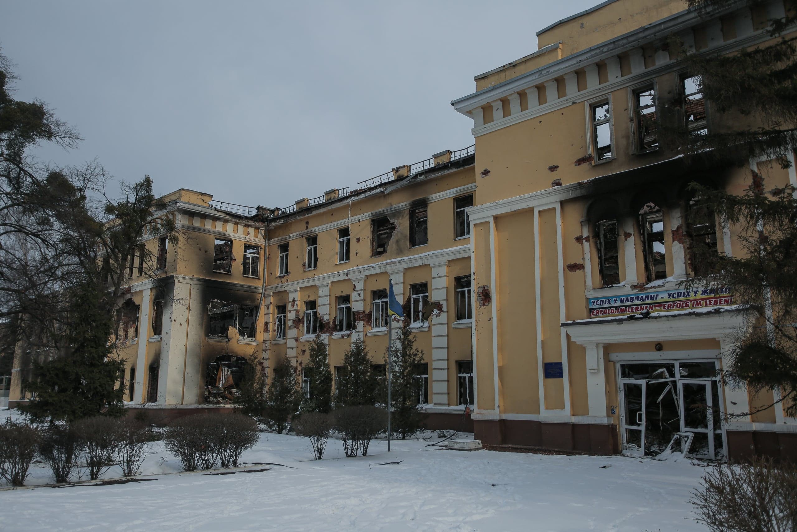 Поврежденное желтое здание учебного заведения на фоне снега. Фасад и выбитые окна покрыты следами обстрелов и копотью. На баннере над входом текст: "УСПІХ У НАВЧАННІ – УСПІХ У ЖИТТІ. ERFOLG IM LERNEN – ERFOLG IM LEBEN".