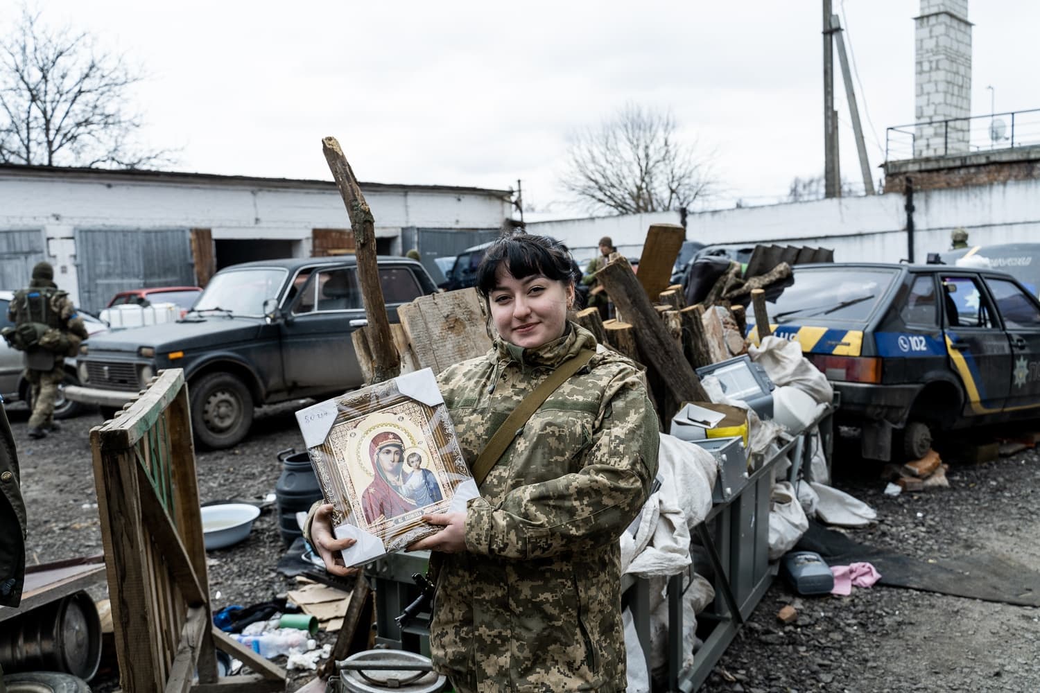 Женщина-военнослужащий в камуфляже улыбается, держа в руках икону Богородицы и Младенца. На фоне замусоренный двор, бревна и автомобиль с полицейской надписью и номером "102".