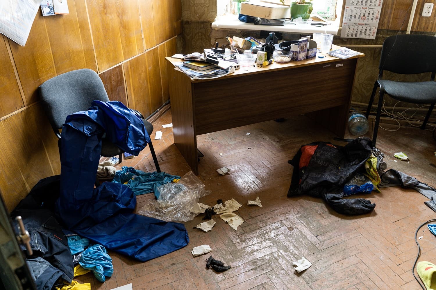 Сильно захламленное офисное помещение с деревянными панелями. Стол завален предметами и бумагами; на паркетном полу разбросана синяя и черная одежда, мусор и обрывки бумаги.