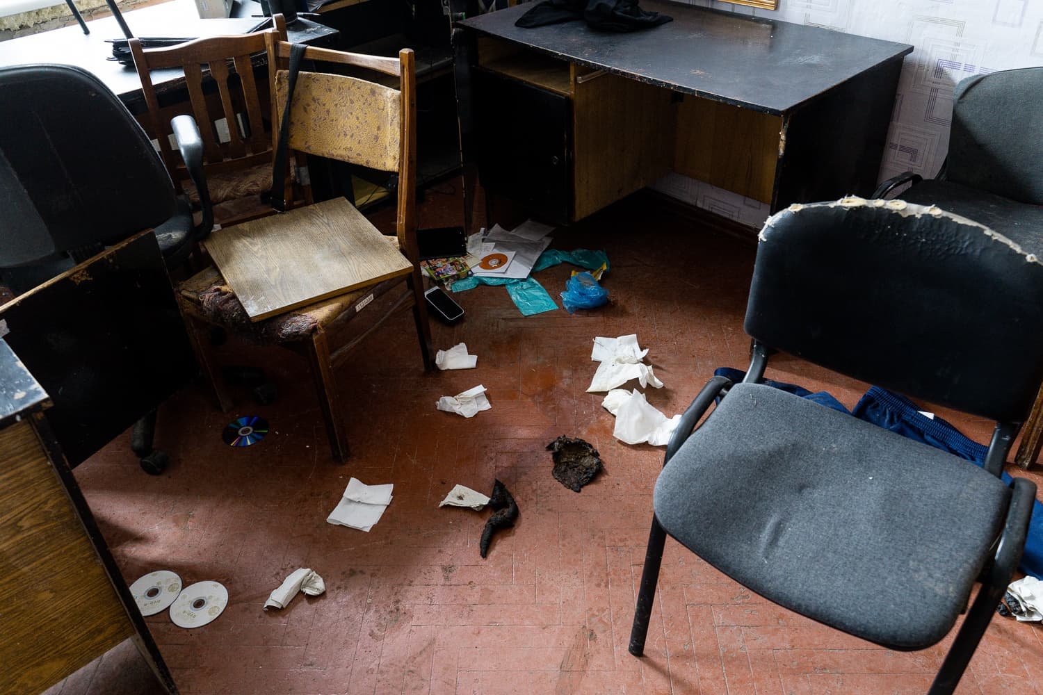 Разгромленное офисное помещение: на грязном полу разбросаны компакт-диски, мусор, скомканные салфетки и темные массы; вокруг стоят старые, поврежденные стулья и стол.