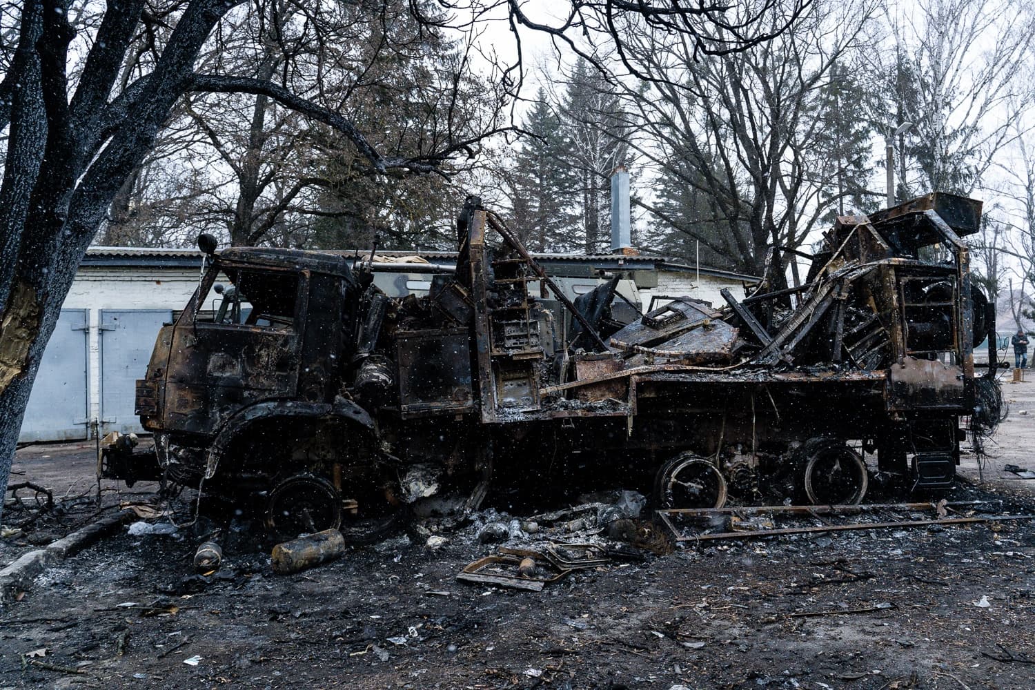 Сгоревший, сильно поврежденный военный грузовик стоит на фоне деревьев и гаражей.