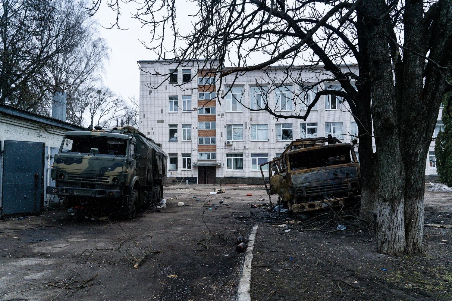 Два грузовика — один камуфляжный, другой сгоревший — стоят перед поврежденным многоэтажным зданием.