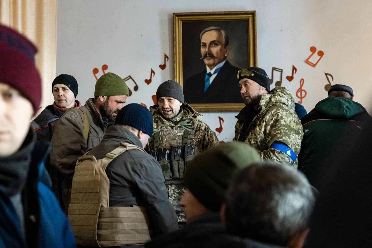 Группа мужчин в военной и повседневной одежде в помещении с портретом усатого мужчины и нотами на стене.