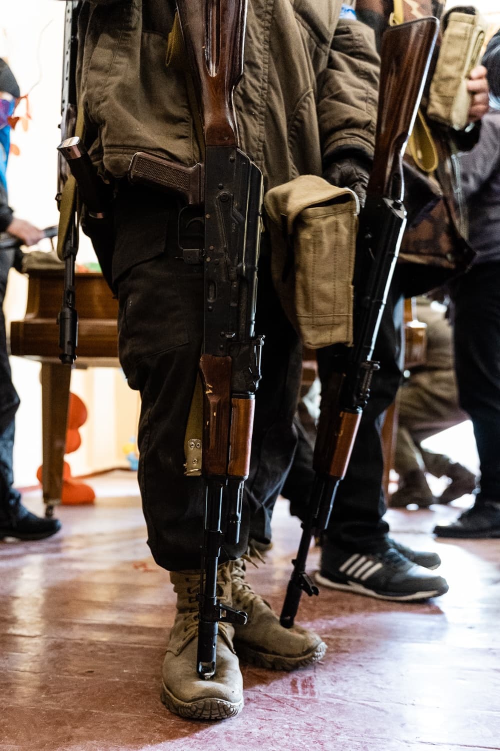 Крупный план нижних частей тел двух человек с автоматами Калашникова (АК) с деревянными прикладами. Люди стоят в помещении в тактической одежде и обуви.