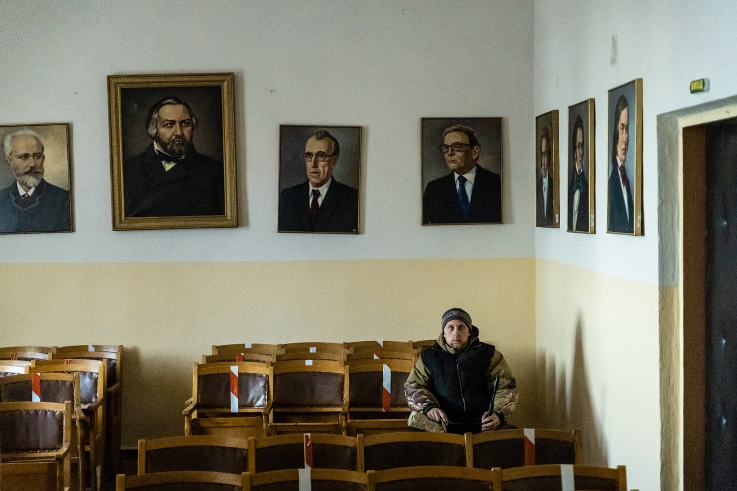 Мужчина в зимней одежде сидит среди рядов деревянных стульев в пустой аудитории. На стене позади него висят несколько портретов известных людей, в том числе П.И. Чайковского.