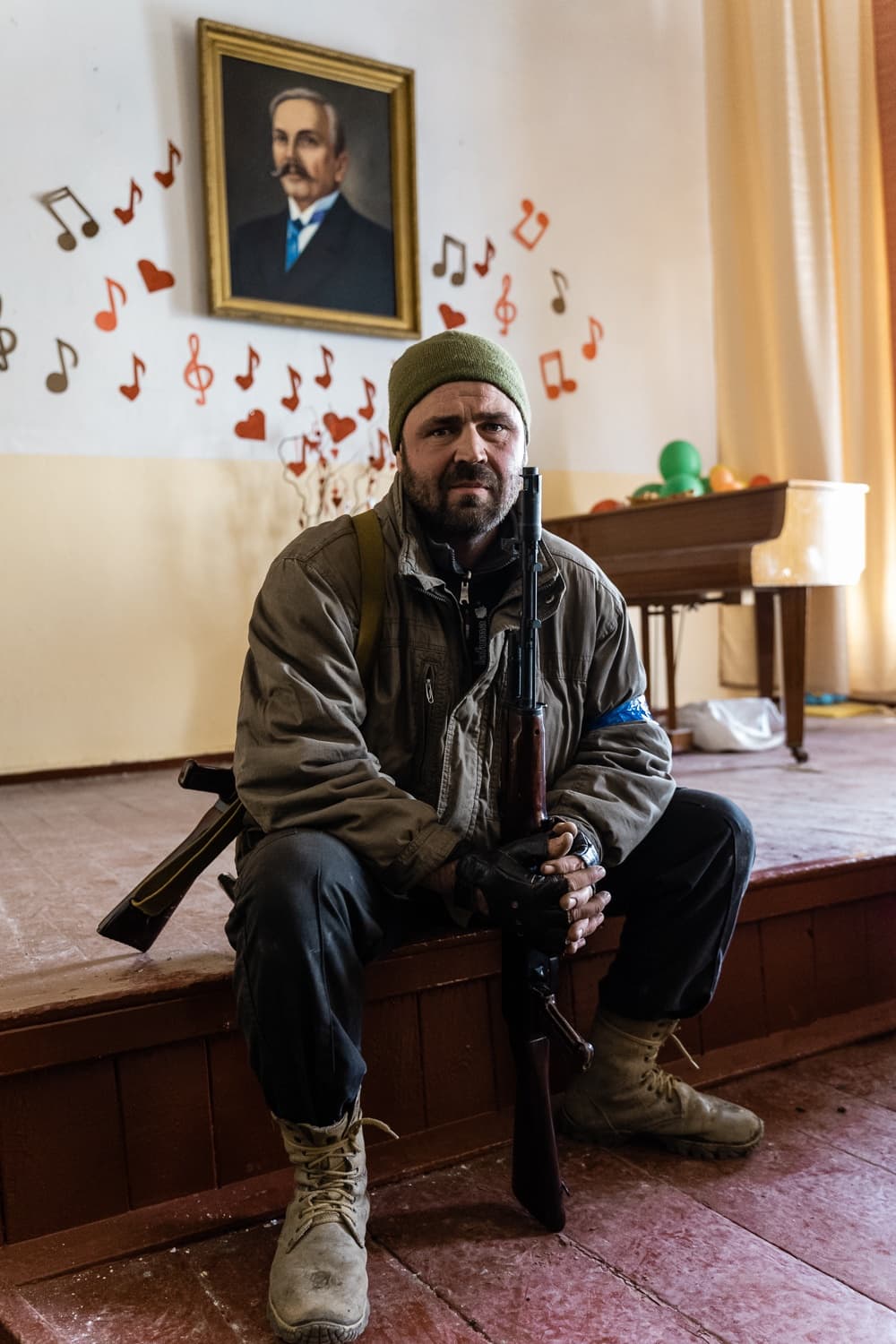 Солдат с винтовкой сидит на краю сцены, украшенной бумажными нотами и сердцами. На стене висит портрет, справа стоит пианино.