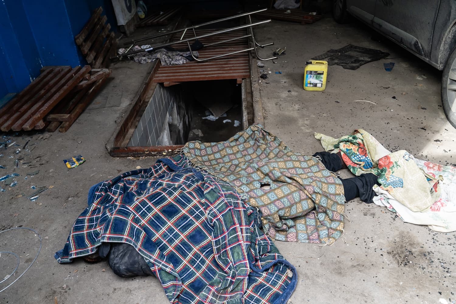 Тело, частично накрытое одеялами, лежит на полу гаража или автосервиса рядом с открытой смотровой ямой и битым стеклом. Видны ноги человека и канистра.