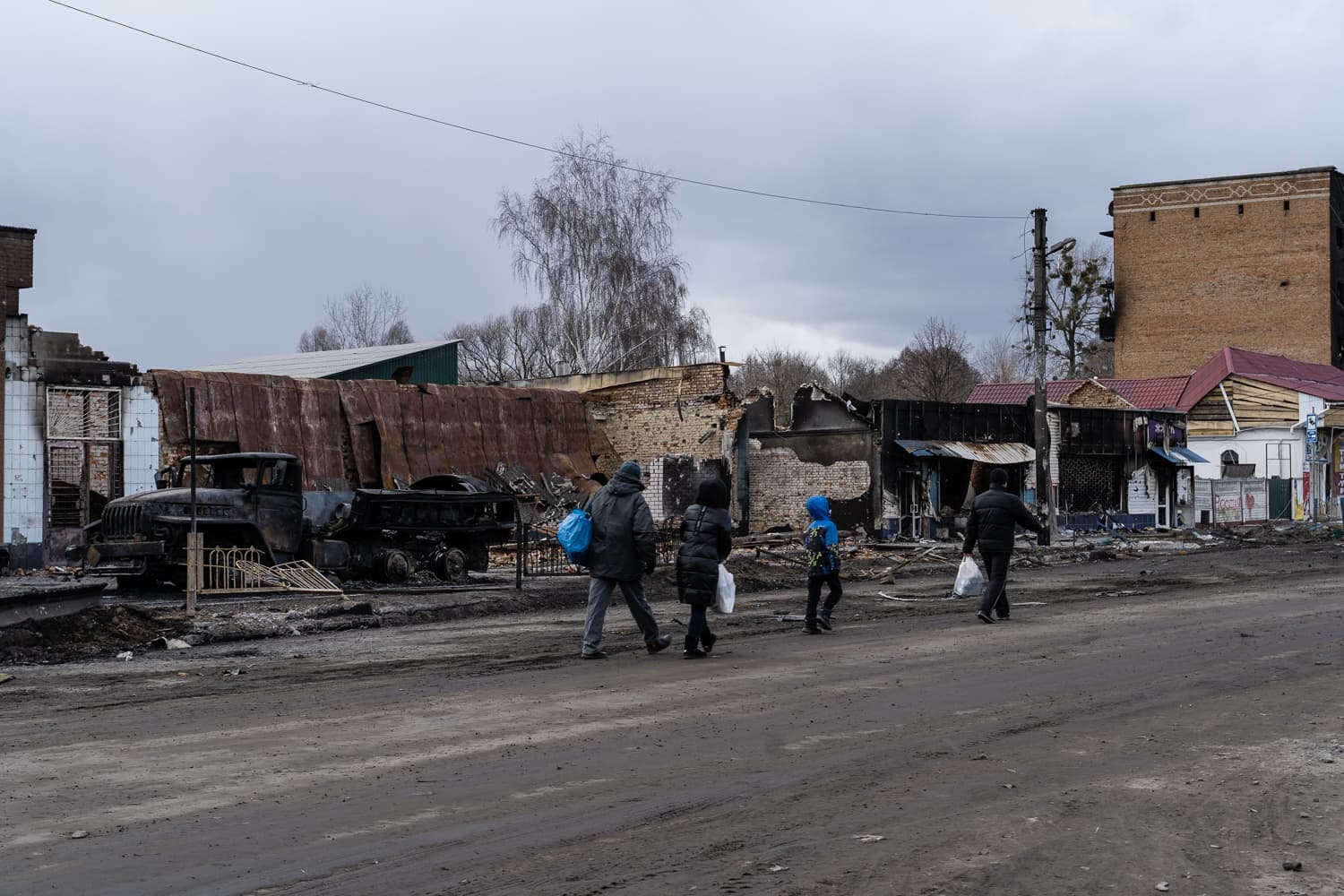 Улица с руинами сгоревших зданий и обгоревшим грузовиком. Четверо людей идут по грязной дороге на фоне серого неба.