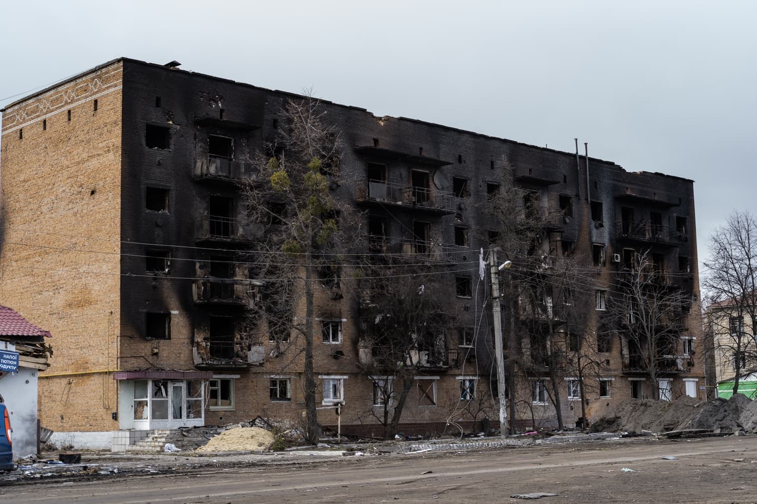Пятиэтажный жилой дом с закопченными черными стенами и пустыми окнами, поврежденный в результате пожара или обстрела. Фасад слева – кирпичный. На земле строительный мусор и грязь. Виден текст: "НАПОЇ ТЮТЮН ПРОДУКТИ".