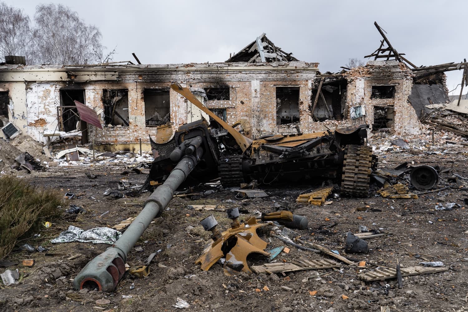 Уничтоженный, сгоревший танк со оторванной башней и пушкой на фоне разрушенного кирпичного здания.