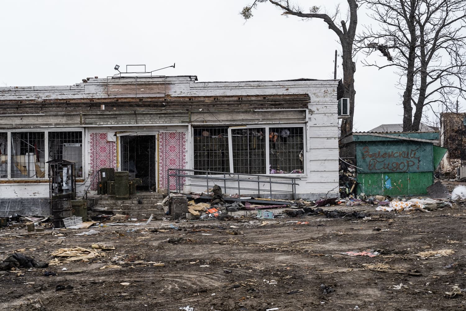 Разрушенный фасад здания с зарешеченными окнами и мусором на грязной земле. Справа зеленый киоск с граффити: "Заленский пидор!".