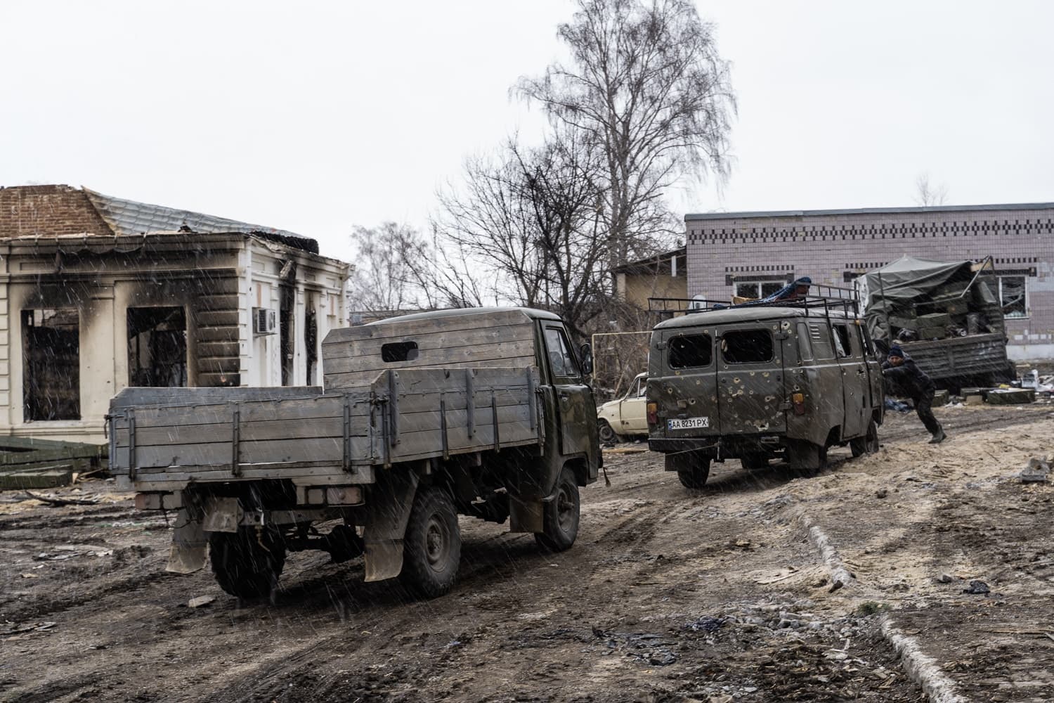 Грязная улица с разрушенными зданиями. Военный грузовик и простреленный микроавтобус ("AA 8231 PX"). Человек толкает микроавтобус. Идет снег.