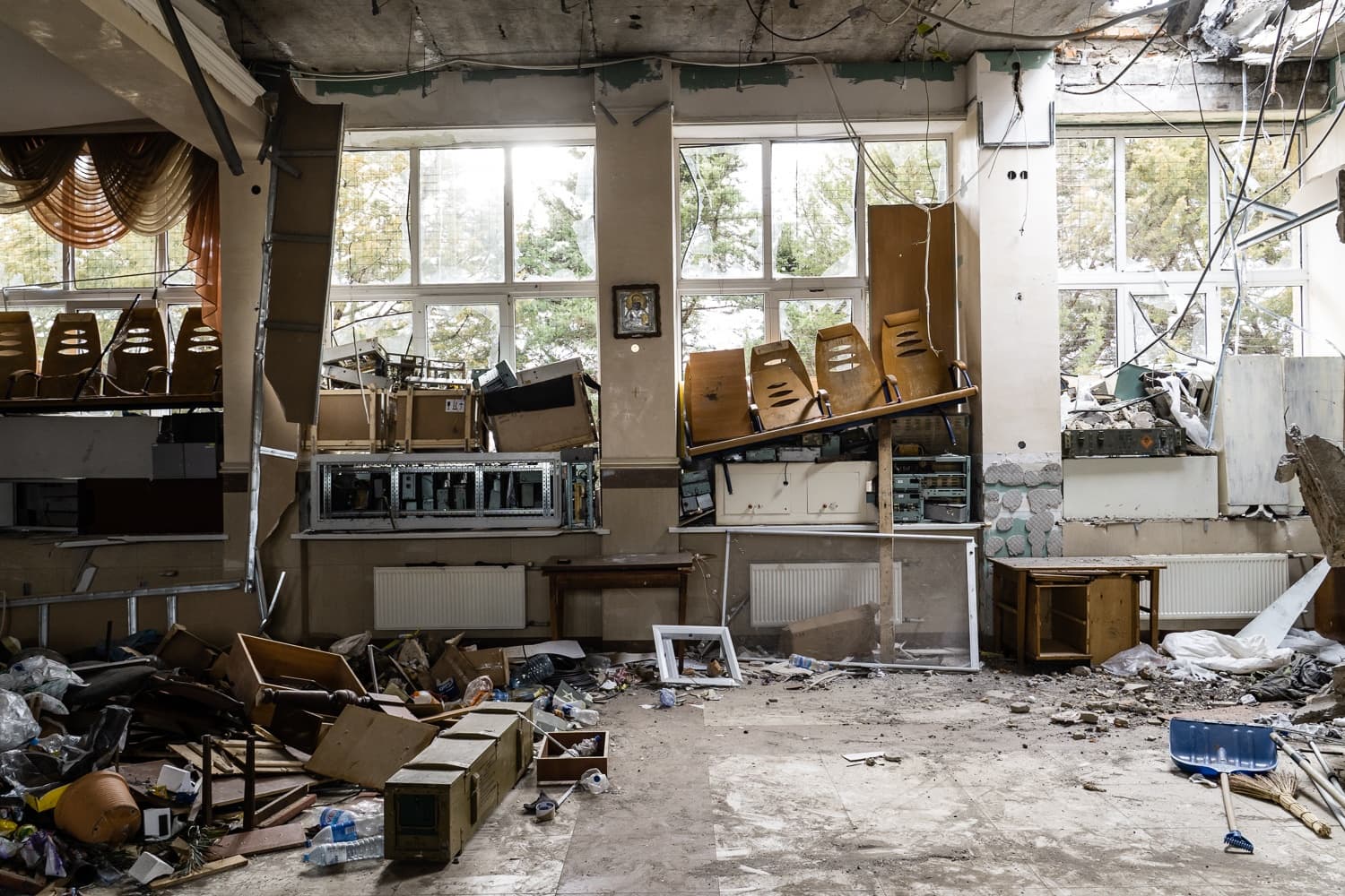 Разрушенный интерьер помещения: разбитые окна, заваленный обломками пол. Поврежденные стены, свисающие провода, сваленные в кучу стулья и ящики.
