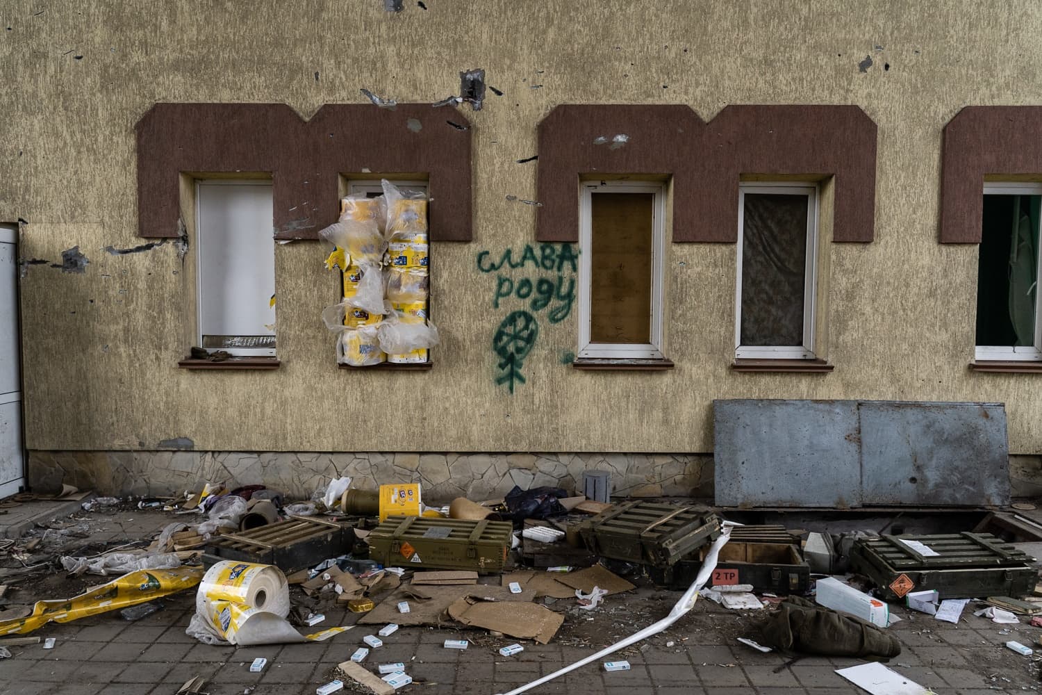 Поврежденная стена здания с граффити «СЛАВА РОДУ» и символом. В одном окне стопки товара. На земле разбросаны мусор и деревянные ящики для боеприпасов.