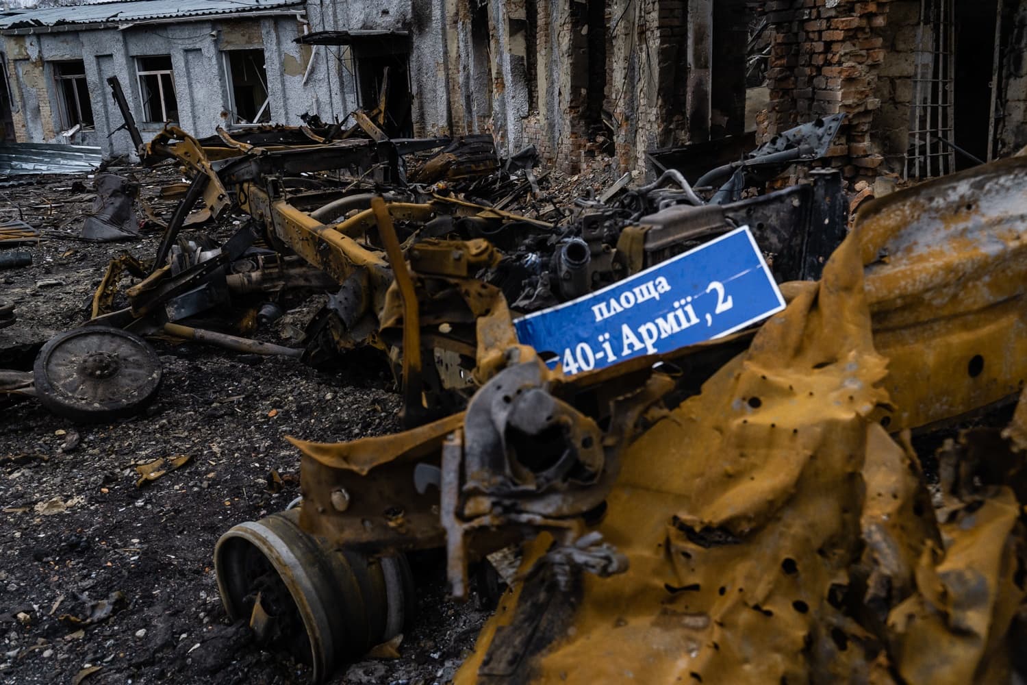 Остатки сгоревшего автомобиля и дорожный знак «Площа 40-ї Армії, 2» на фоне разрушенного здания.