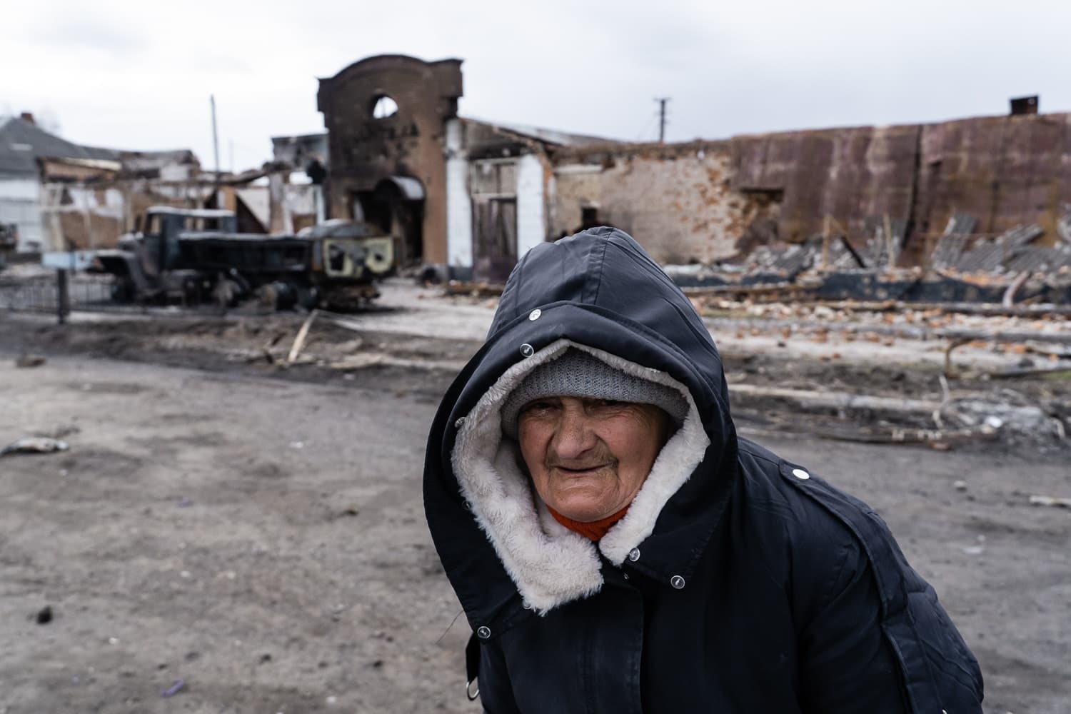 Пожилая женщина в капюшоне и серой шапке на фоне разрушенных войной зданий и сгоревшего грузовика.