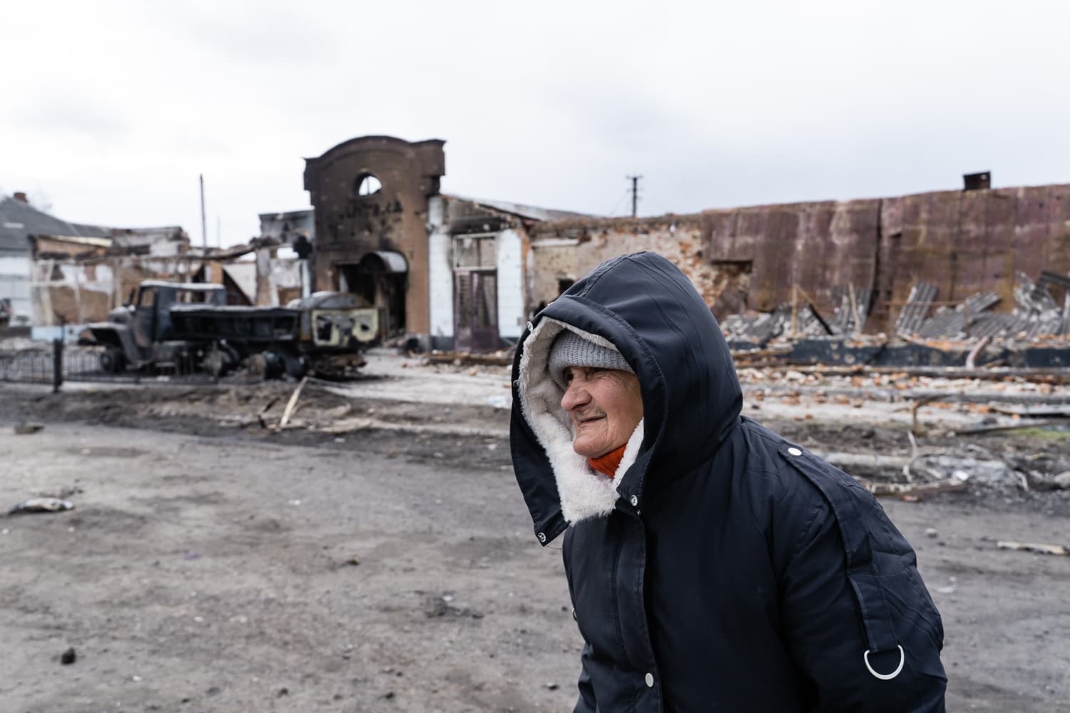 Пожилая женщина в капюшоне стоит на фоне разрушенных войной зданий и сгоревшего грузовика.