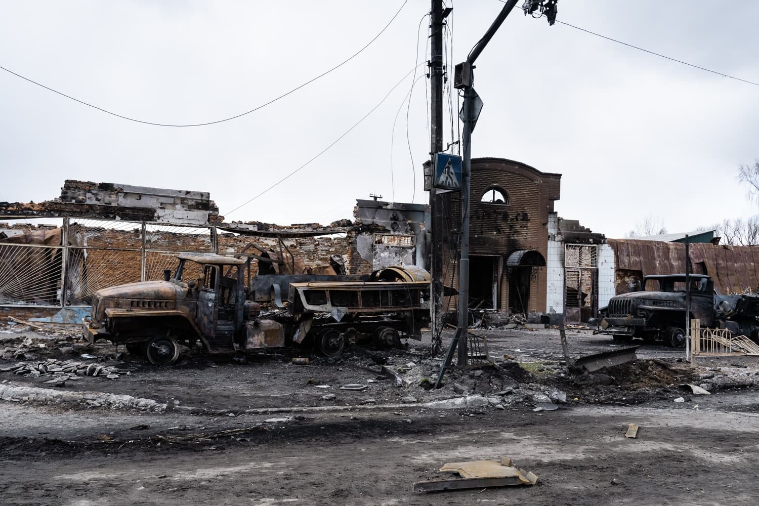 Обугленные остовы двух грузовиков на грязной улице среди разрушенных и почерневших зданий.