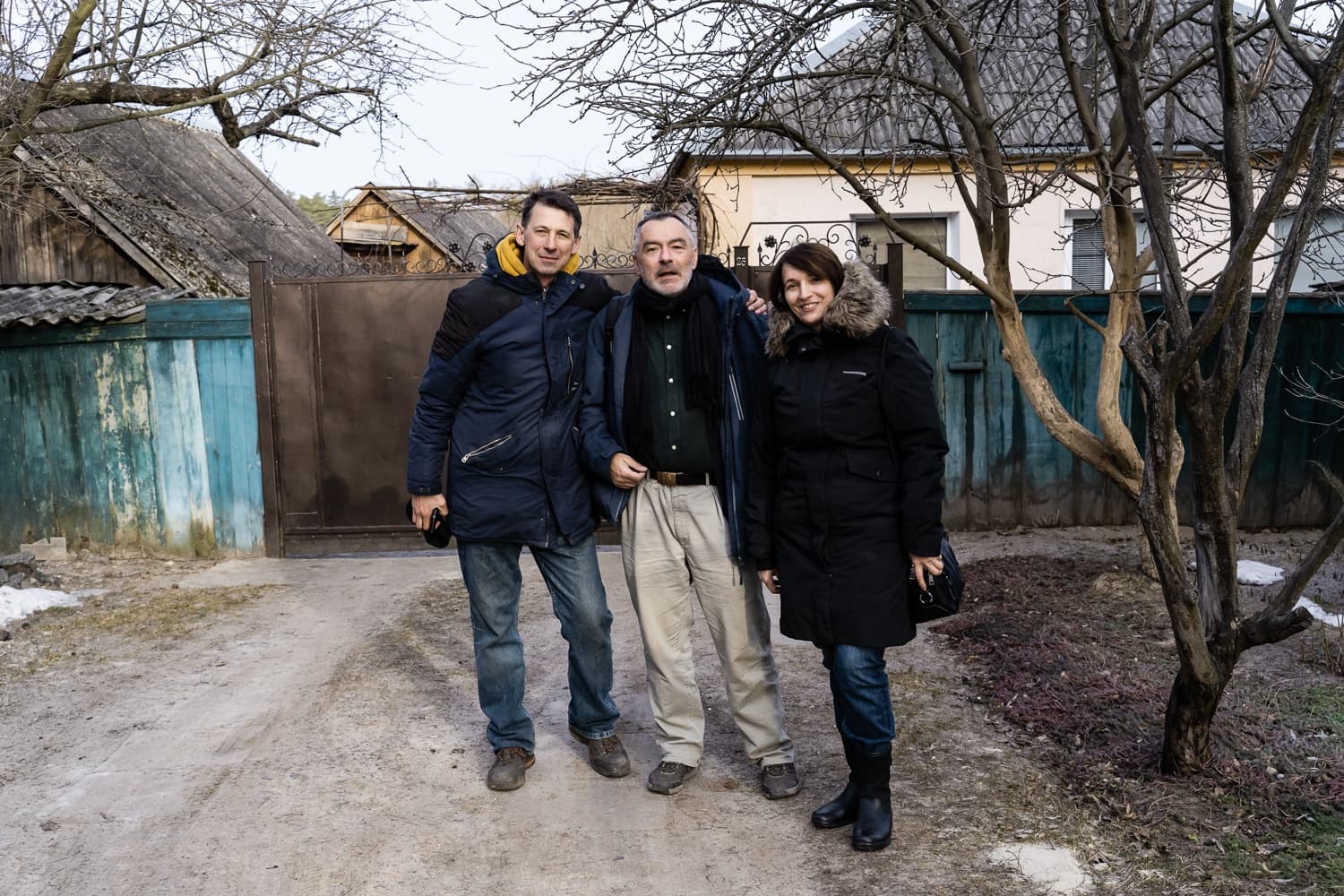 Групповой портрет троих людей в зимних куртках на фоне сельского двора, забора и построек.