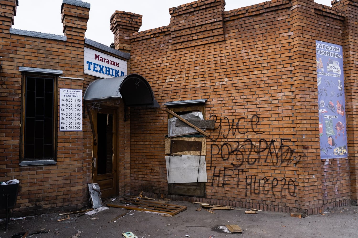 Здание магазина "Техника" с разбитой дверью, заколоченным окном и граффити на стене: "Уже розграб*но нет ничего".