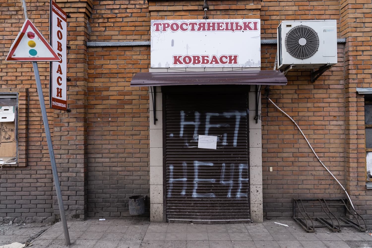Фасад магазина с кирпичной кладкой и вывеской "ТРОСТЯНЕЦЬКІ КОВБАСИ". На закрытой металлической роллете белое граффити "НЕТ" и "НЕЧА". Слева виден дорожный знак.