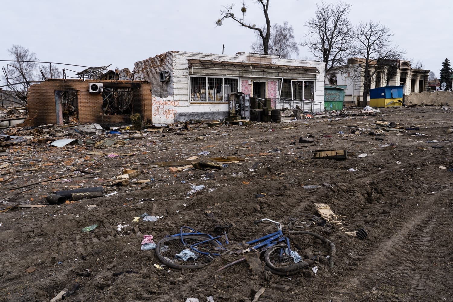 Разрушенные войной здания и грязный пустырь, заваленный обломками. На переднем плане лежит синий велосипед. На стене одного из зданий граффити: "Зеленський Я".